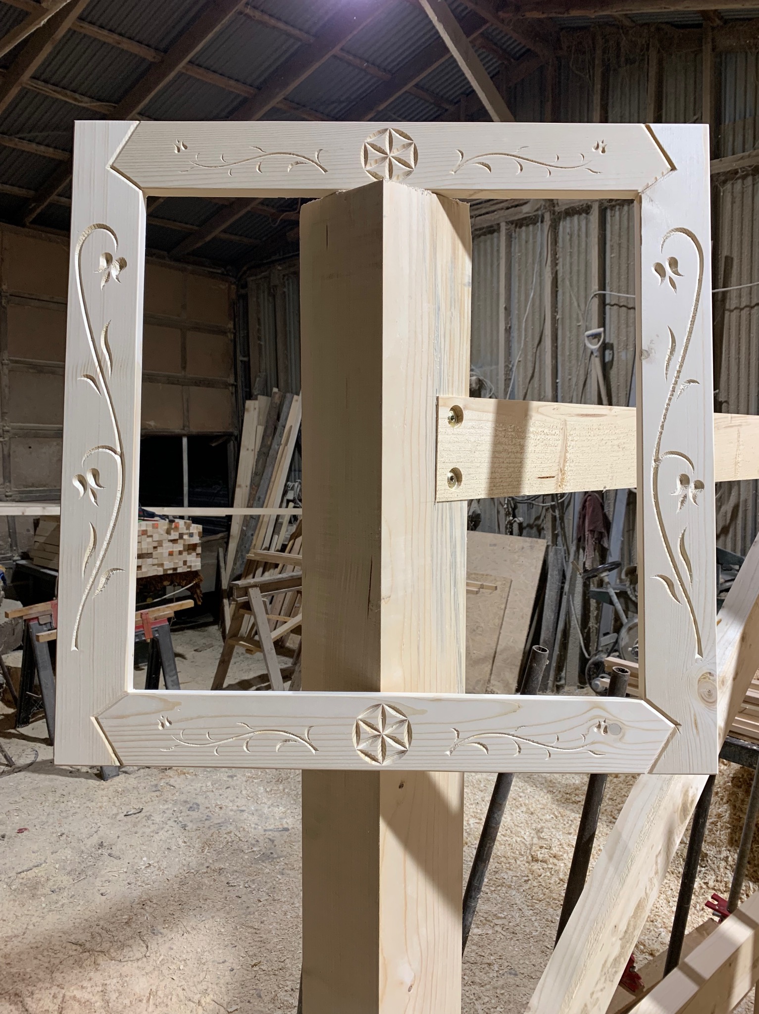 Surowa, drewniana rama z wyciętymi wzorami kwiatowymi i geometrycznymi, umieszczona na drewnianym słupie w warsztacie stolarskim.