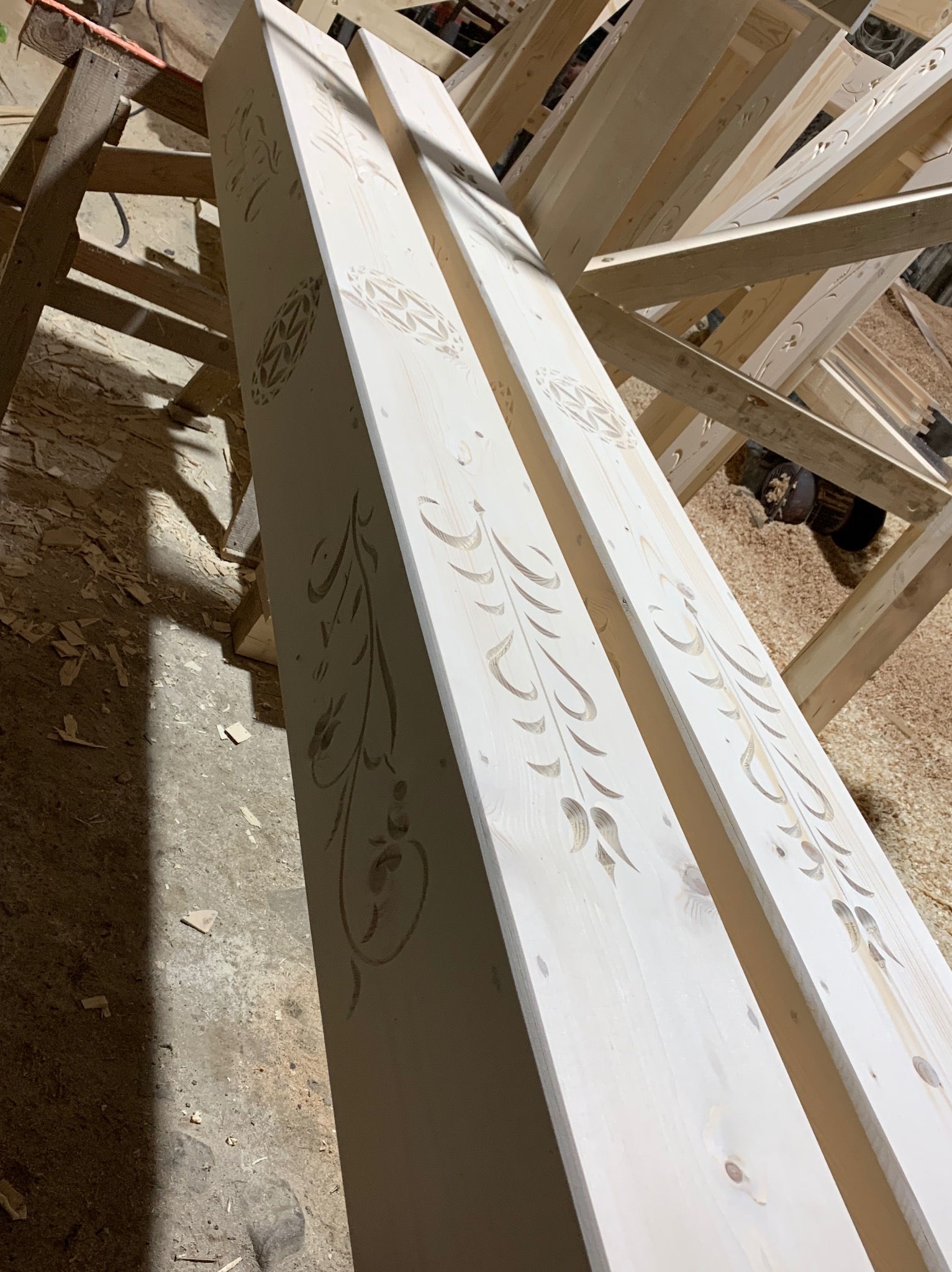 Dwa jasne, drewniane tragarze z wyżłobionymi, dekoracyjnymi wzorami roślinnymi i geometrycznymi, ustawione na drewnianym stojaku w warsztacie.