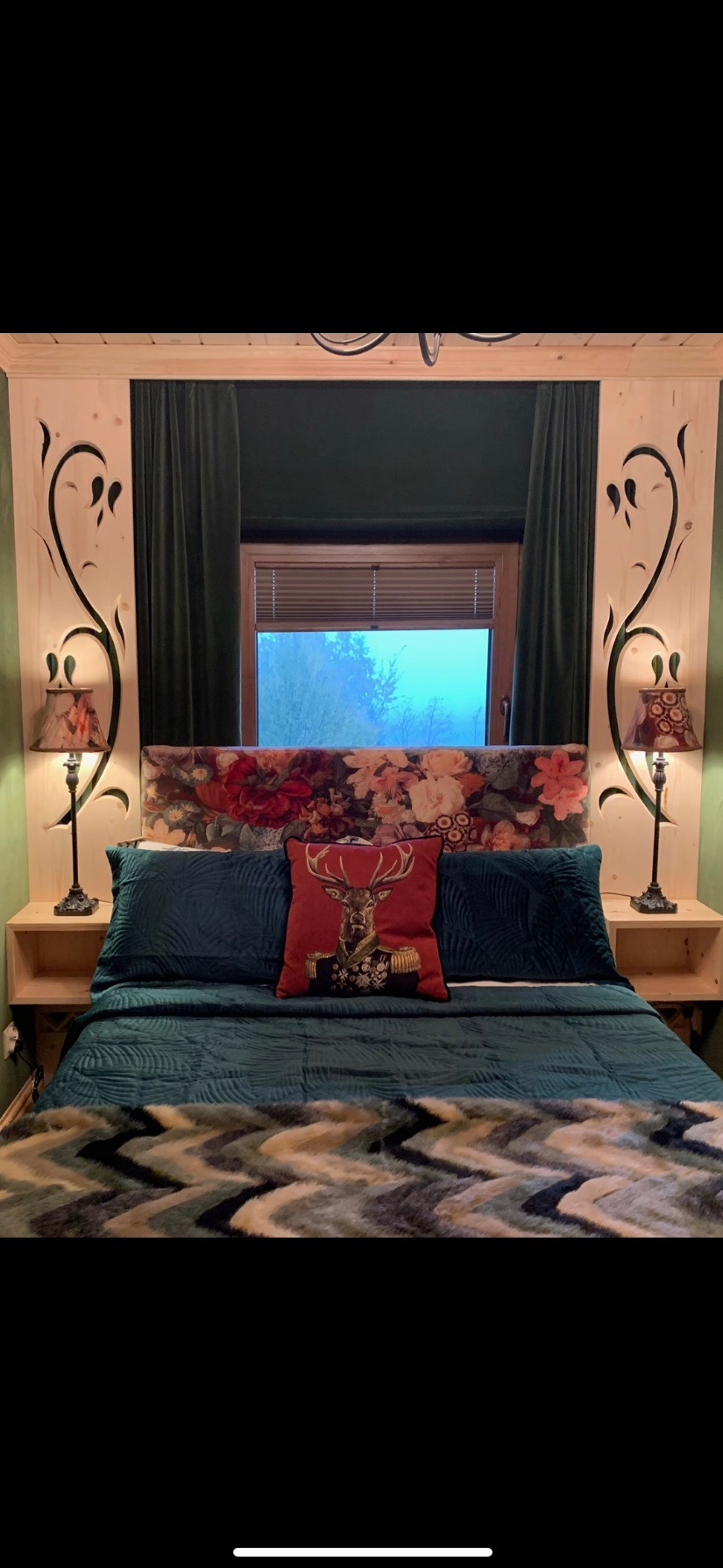 Sypialnia z łóżkiem w stylu rustykalnym, drewniane panele z wyciętymi wzorami roślinnymi po bokach, kwiatowy zagłówek, zielone poduszki i narzuta, czerwona poduszka z wizerunkiem jelenia w mundurze...