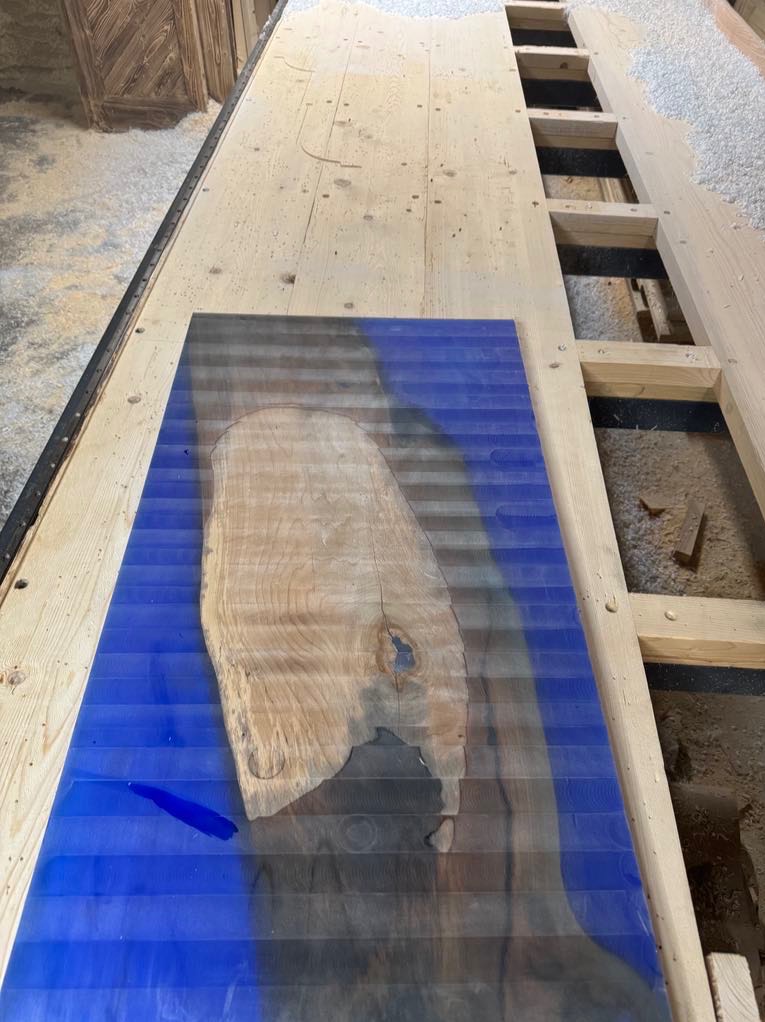 Blat stołu z żywicy epoksydowej w kolorze niebieskim, z wyraźnym usłojeniem drewna, leżący na drewnianym stole warsztatowym z widocznymi wiórami.