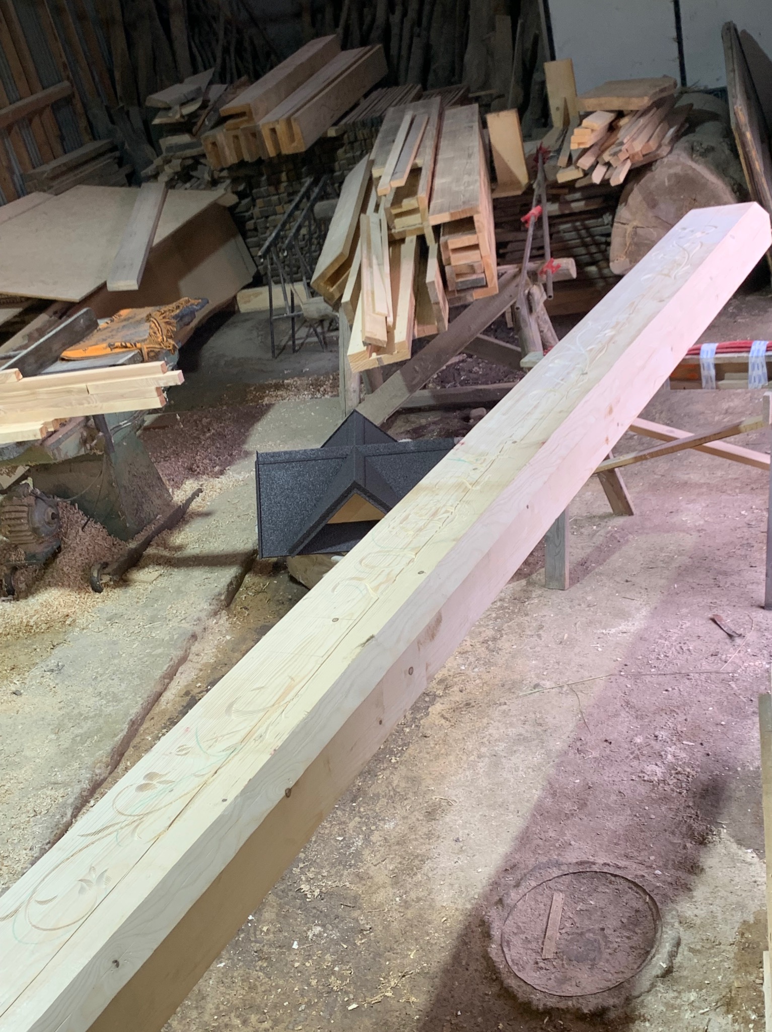 Długa, jasna belka z drewna z delikatnymi, wyżłobionymi wzorami kwiatowymi, leżąca ukośnie w warsztacie, z widocznymi stosami drewna i narzędziami w tle.