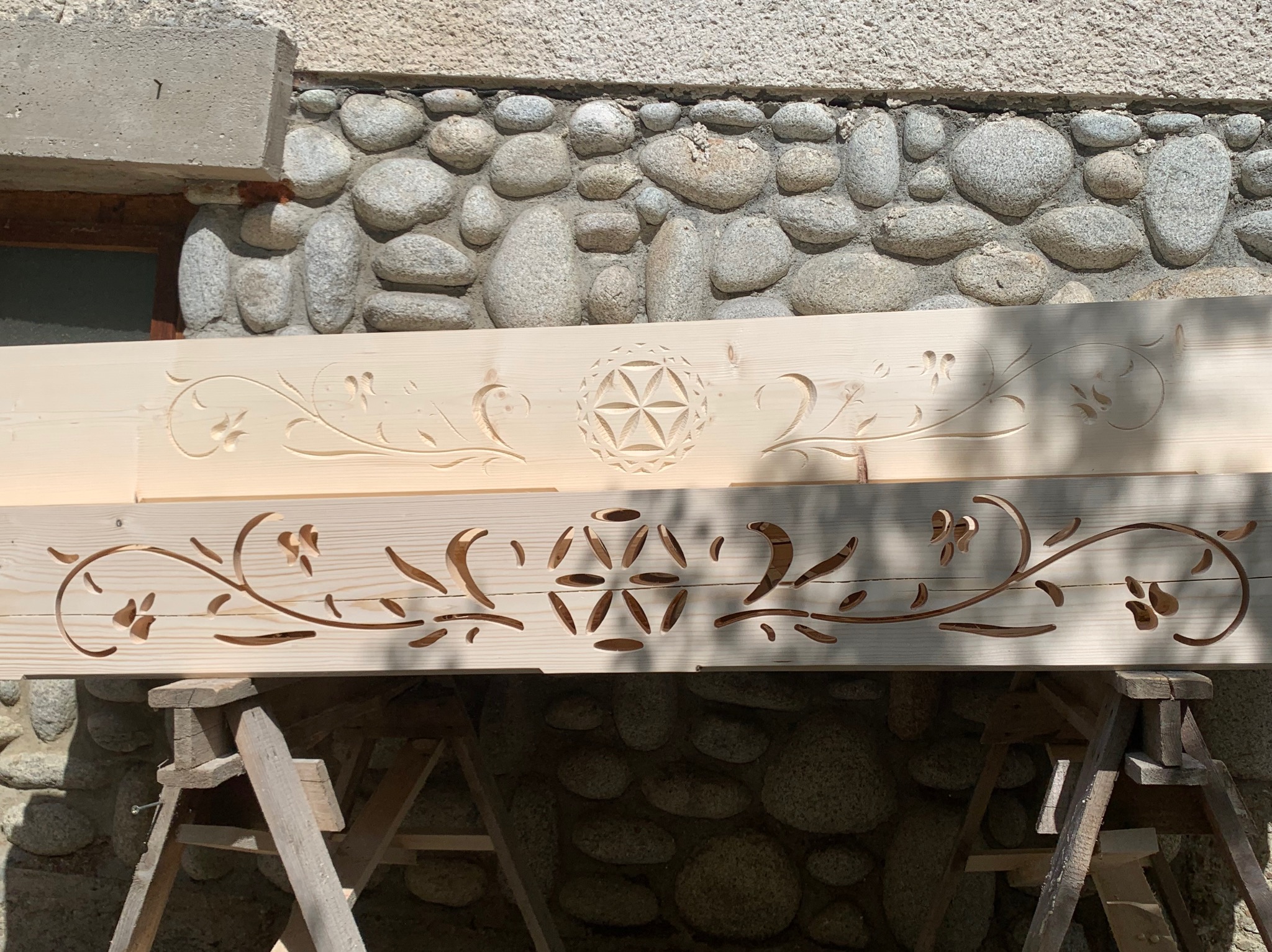 Drewniana belka z wyciętymi wzorami kwiatowymi i geometrycznymi, oparta na drewnianych kozłach, na tle kamiennej ściany.