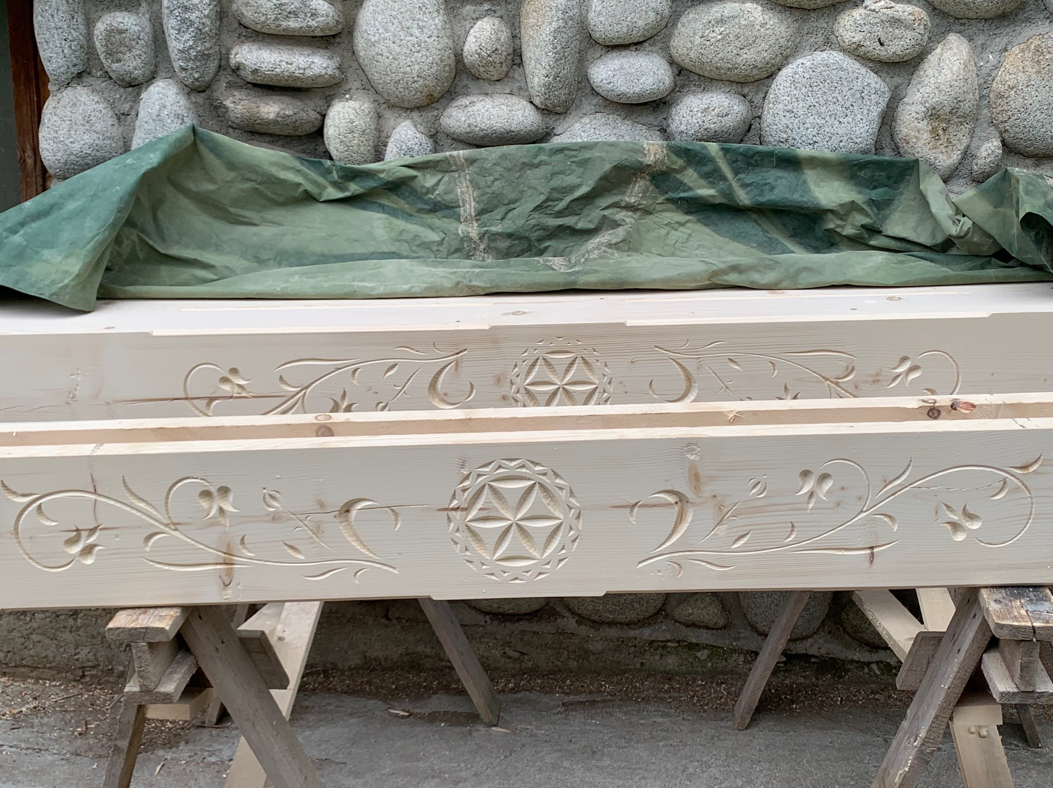 Dwa surowe, jasne drewniane elementy konstrukcyjne z rzeźbionymi wzorami kwiatowymi i geometrycznym motywem rozety, spoczywające na drewnianych kozłach na tle kamiennej ściany i zielonej plandeki.