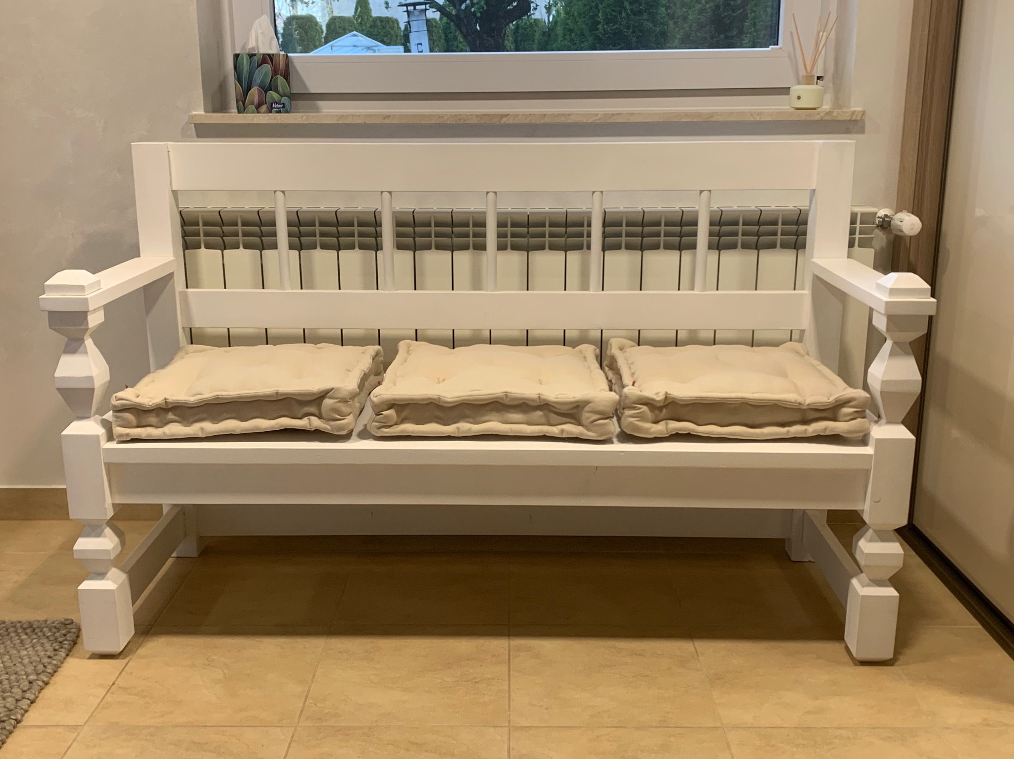 Biała, drewniana ławka z trzema beżowymi poduszkami na siedzisku, ustawiona pod oknem z widokiem na drzewa, z grzejnikiem w tle, na podłodze z płytek ceramicznych.