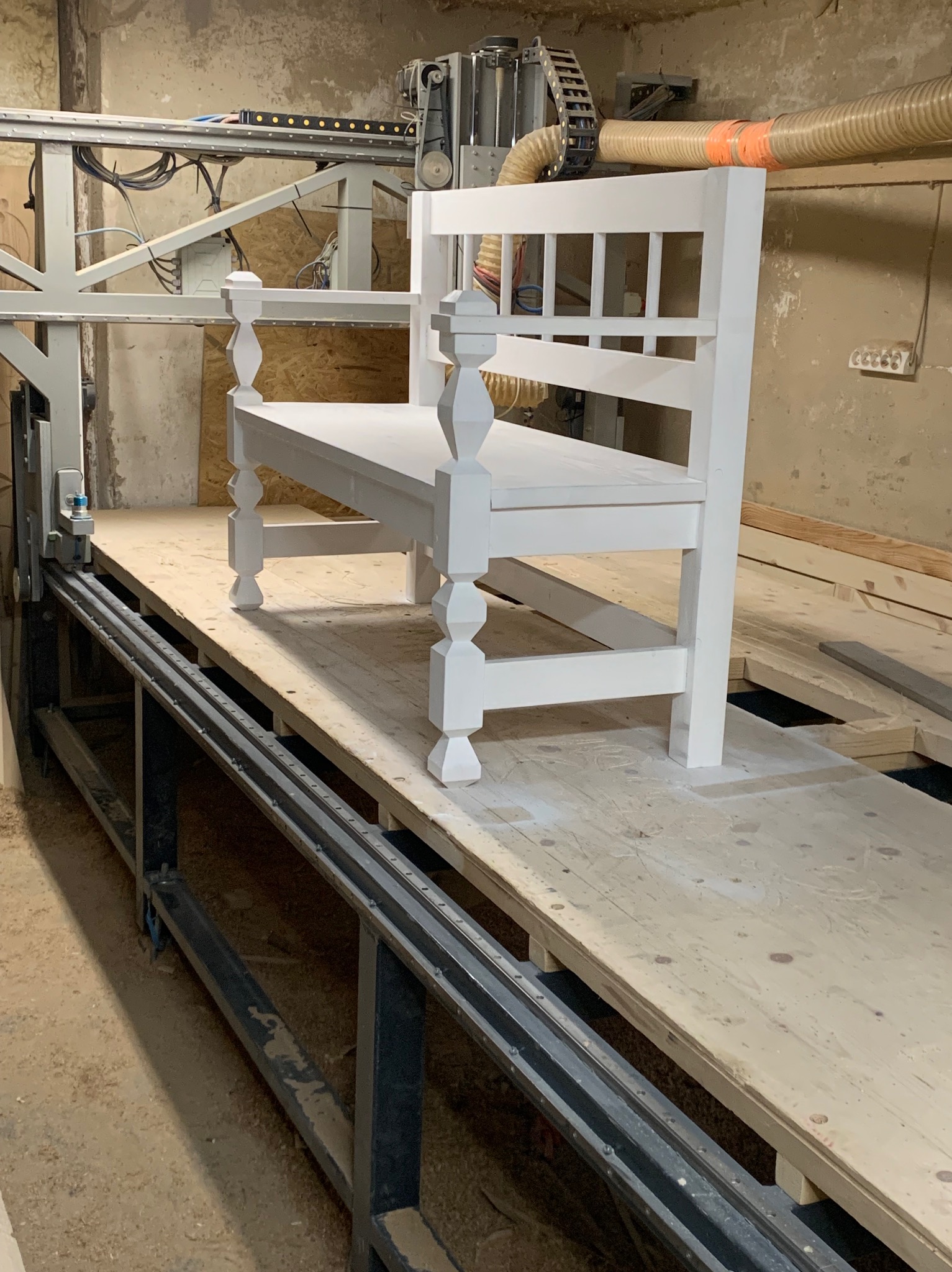 Biała, drewniana ławka z oparciem, częściowo obrobiona, umieszczona na platformie obrabiarki CNC w warsztacie stolarskim.