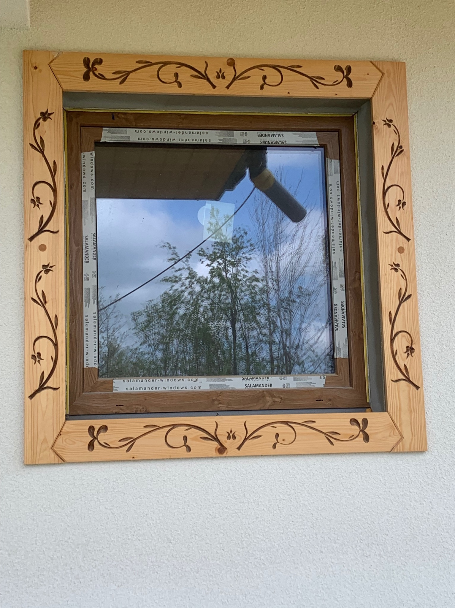 Okno z drewnianą ramą zdobioną motywami roślinnymi, widok na ogród przez szybę.