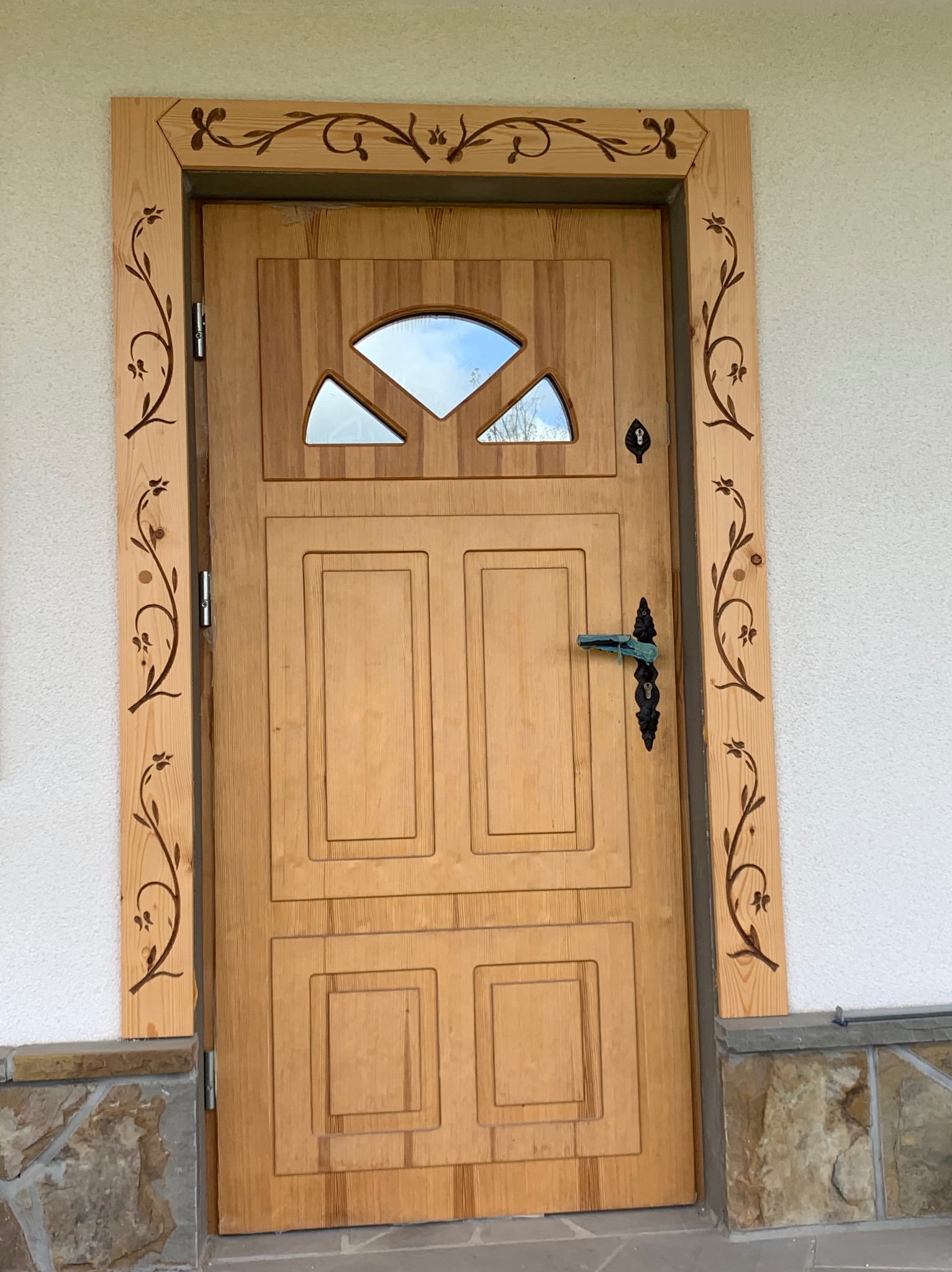 Drewniane drzwi zewnętrzne z ozdobną ramą i szybą w górnej części, wyposażone w czarną klamkę i zamek, zamontowane w białej ścianie z kamienną podmurówką.