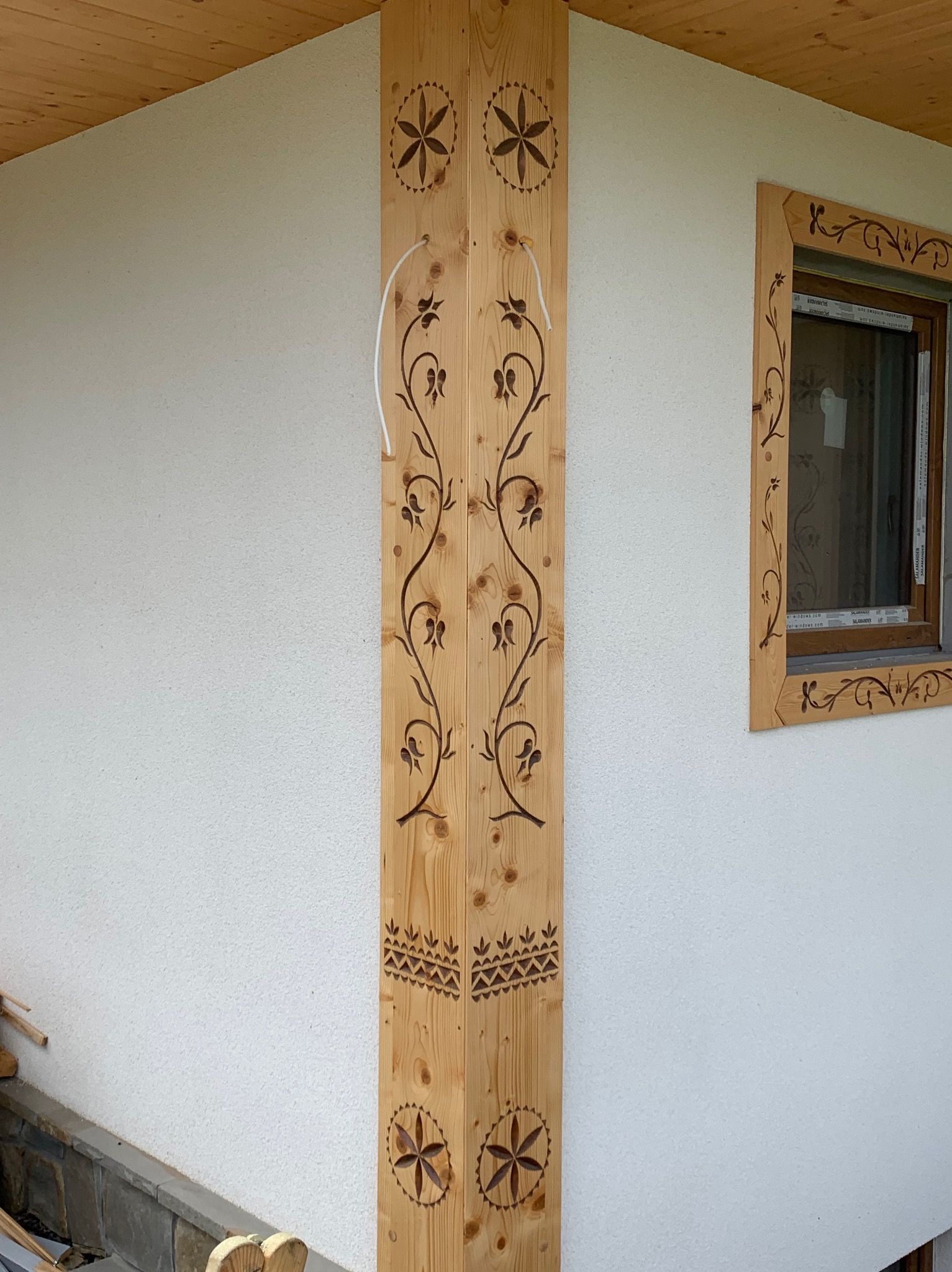 Drewniana kolumna narożna z dekoracyjnymi, wypalanymi wzorami kwiatowymi i geometrycznymi, dopasowana do zdobień ramy okiennej na tle białego, teksturowanego muru.