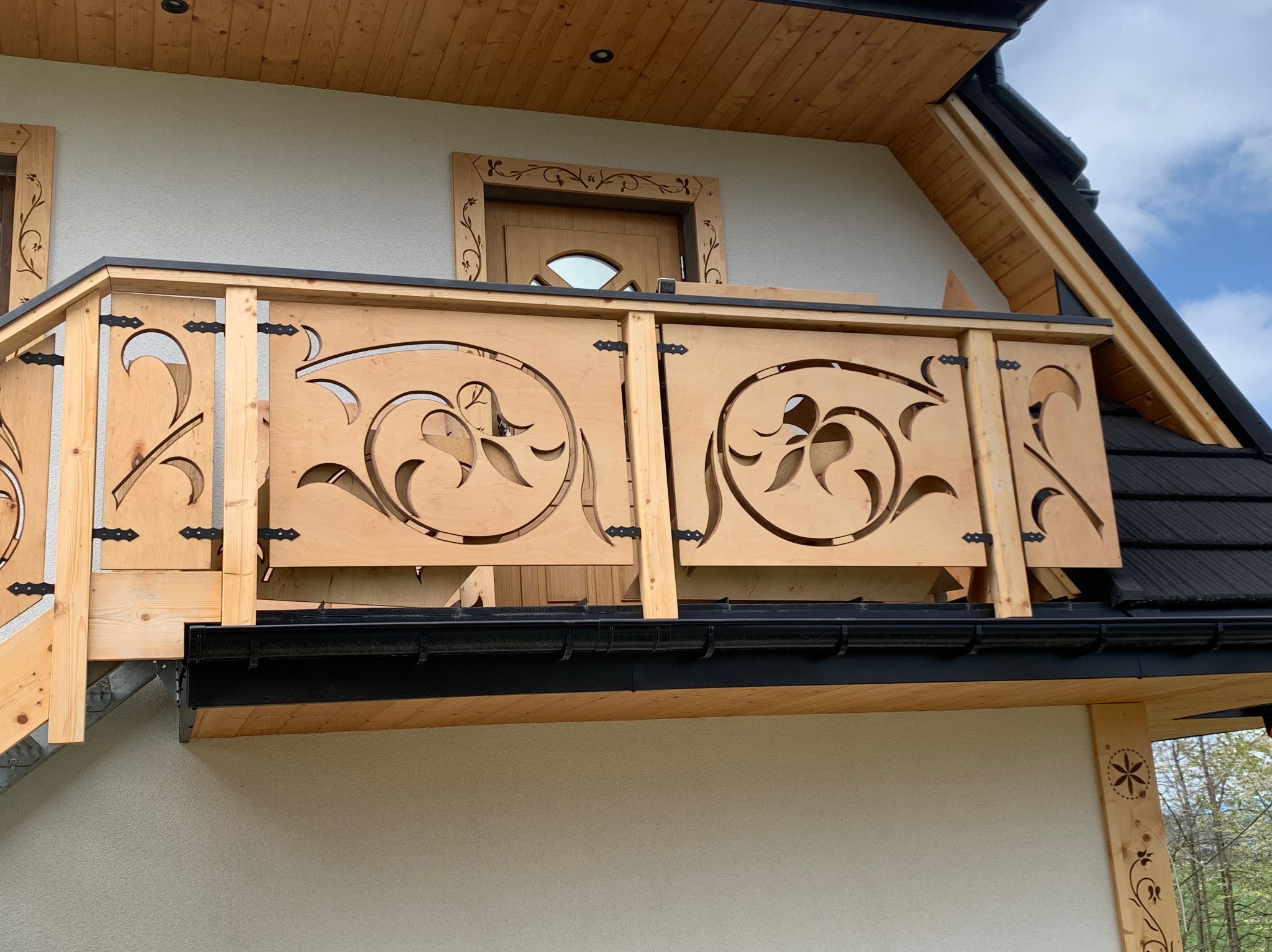 Balustrada balkonowa z jasnego drewna z wyciętymi motywami roślinnymi, czarne okucia zawiasowe, w tle fragment elewacji budynku z drewnianą stolarką okienną z motywami kwiatowymi.