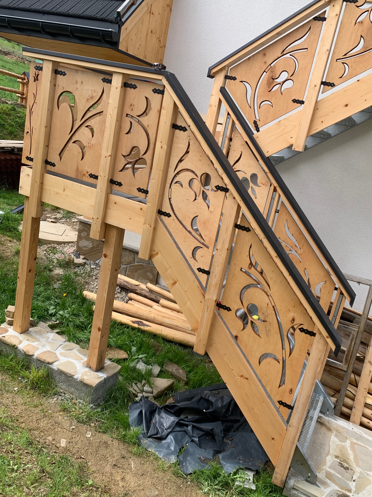 Zewnętrzne schody drewniane z ozdobnymi wycięciami w balustradach, czarne elementy wykończeniowe, częściowo wykonane, widoczne materiały budowlane obok.