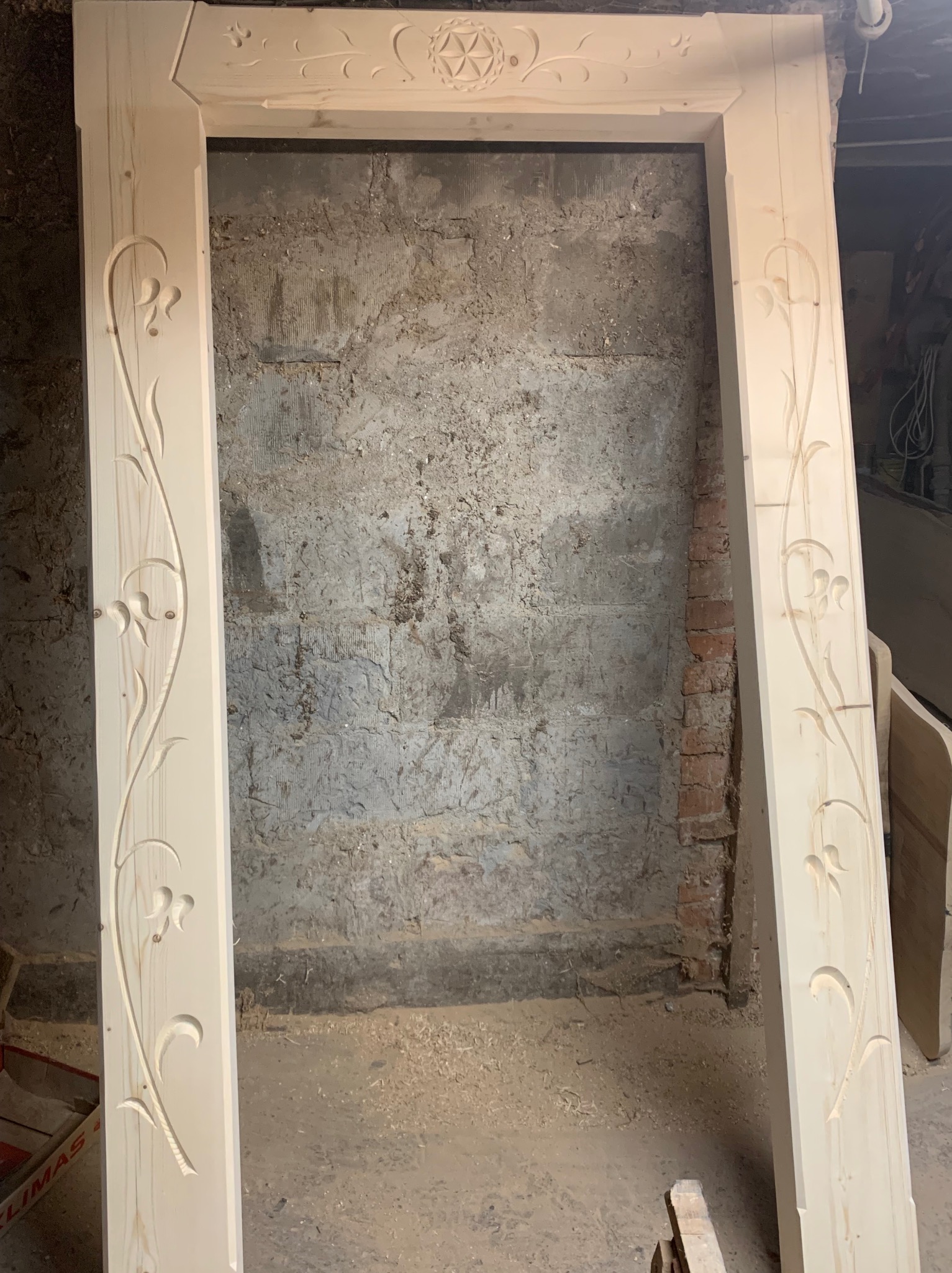 Surowa, drewniana rama drzwiowa z wyżłobionymi motywami kwiatowymi, oparta o szarą, surową ścianę w trakcie obróbki w warsztacie.