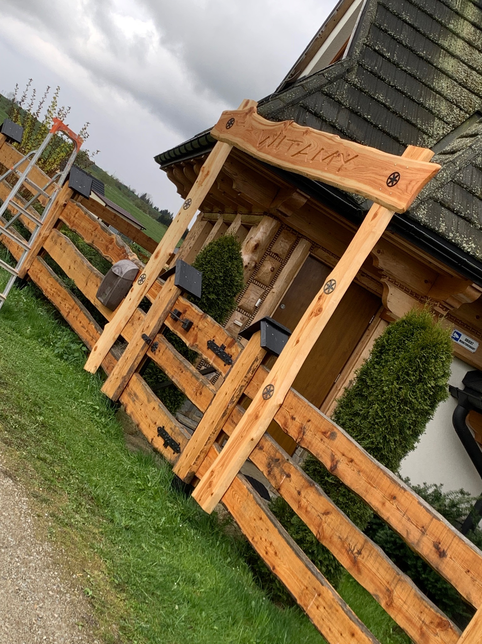 Nowe, jasne ogrodzenie z drewna w stylu góralskim z ozdobnymi elementami i napisem 'WITAM' nad wejściem, częściowo zasłaniające budynek, z drabinką opartą o bok ogrodzenia.