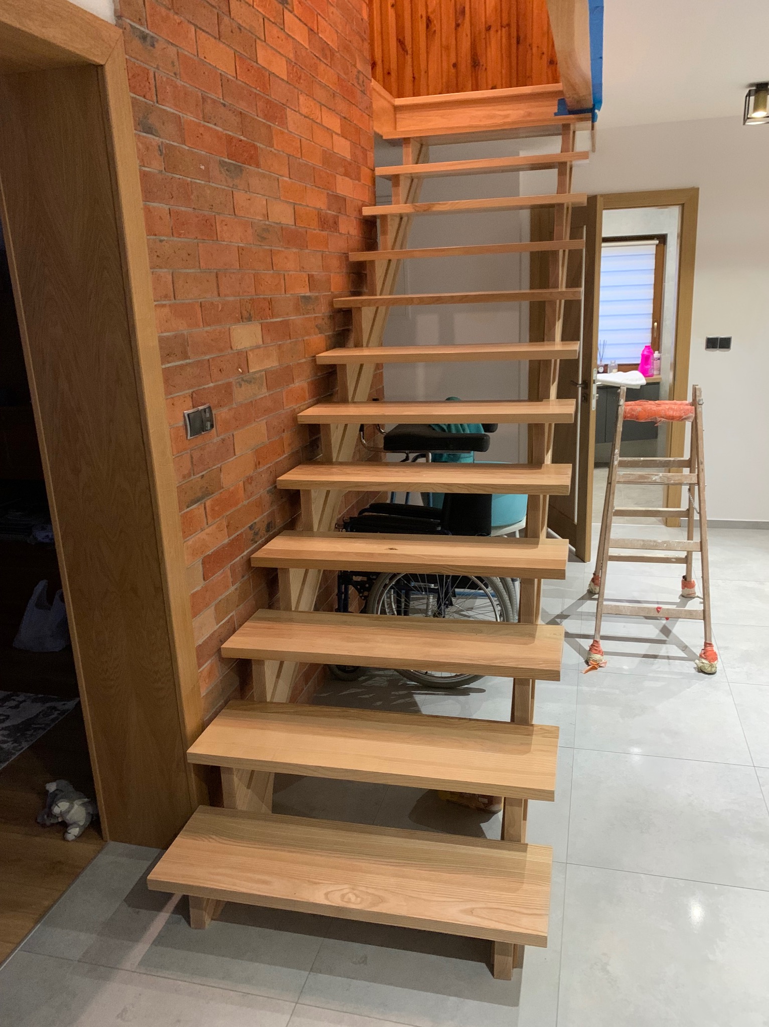 Surowe, drewniane stopnie schodów przy ceglanej ścianie, widoczna drabina malarska i wózek inwalidzki w tle.