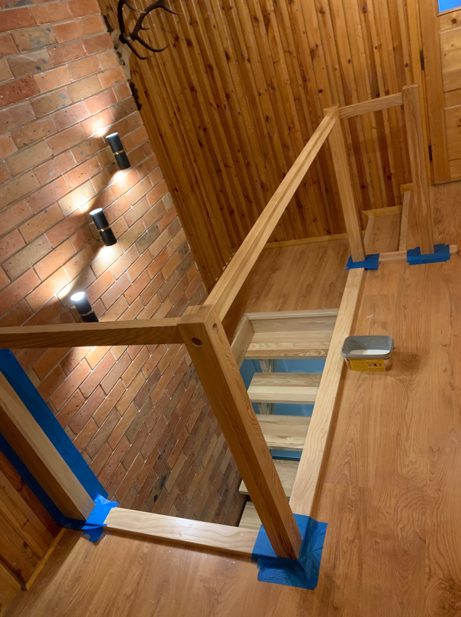 Wnętrze z drewnianymi schodami w trakcie montażu, zabezpieczone niebieską taśmą malarską, widoczna puszka z preparatem do drewna, ściana z cegły i drewniana boazeria.