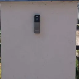 Szary domofon z klawiaturą numeryczną i kamerą zamontowany na kremowym słupku ogrodzeniowym, widok z zewnątrz.