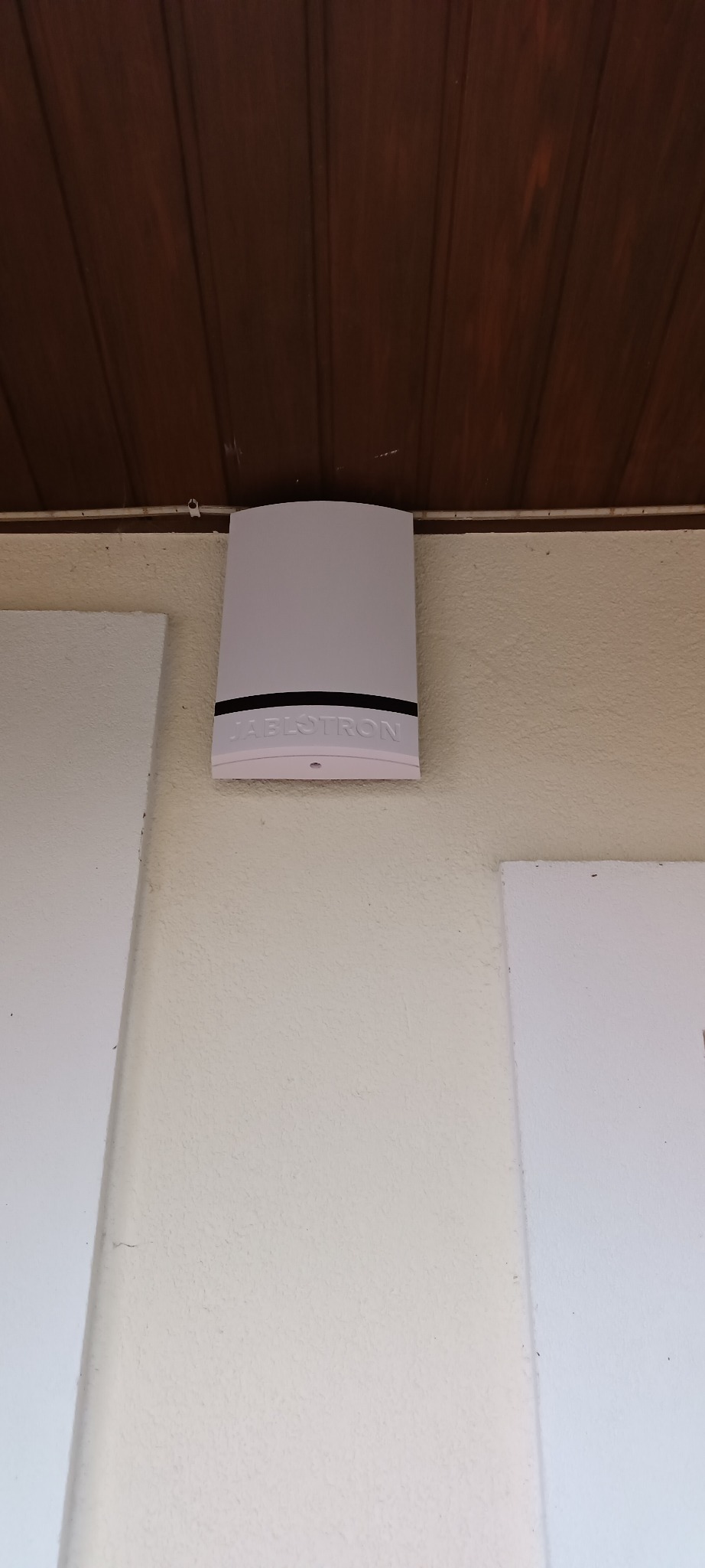 Biała centrala alarmowa Jablotron zamontowana na kremowej ścianie pod drewnianym sufitem.