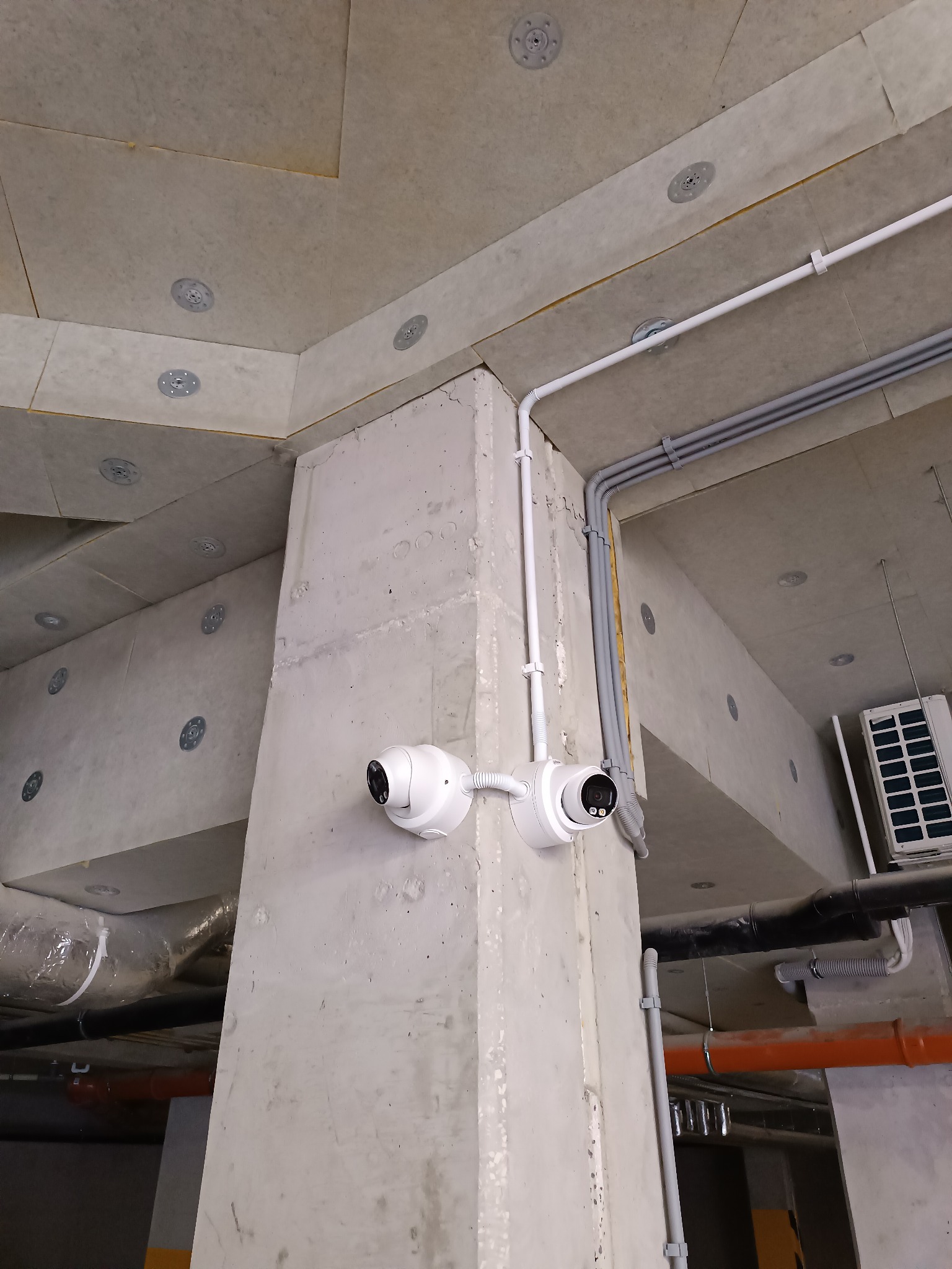 Dwie kamery monitoringu kopułkowe zamontowane na betonowym słupie w garażu podziemnym; widoczne rury instalacyjne i klimatyzator.
