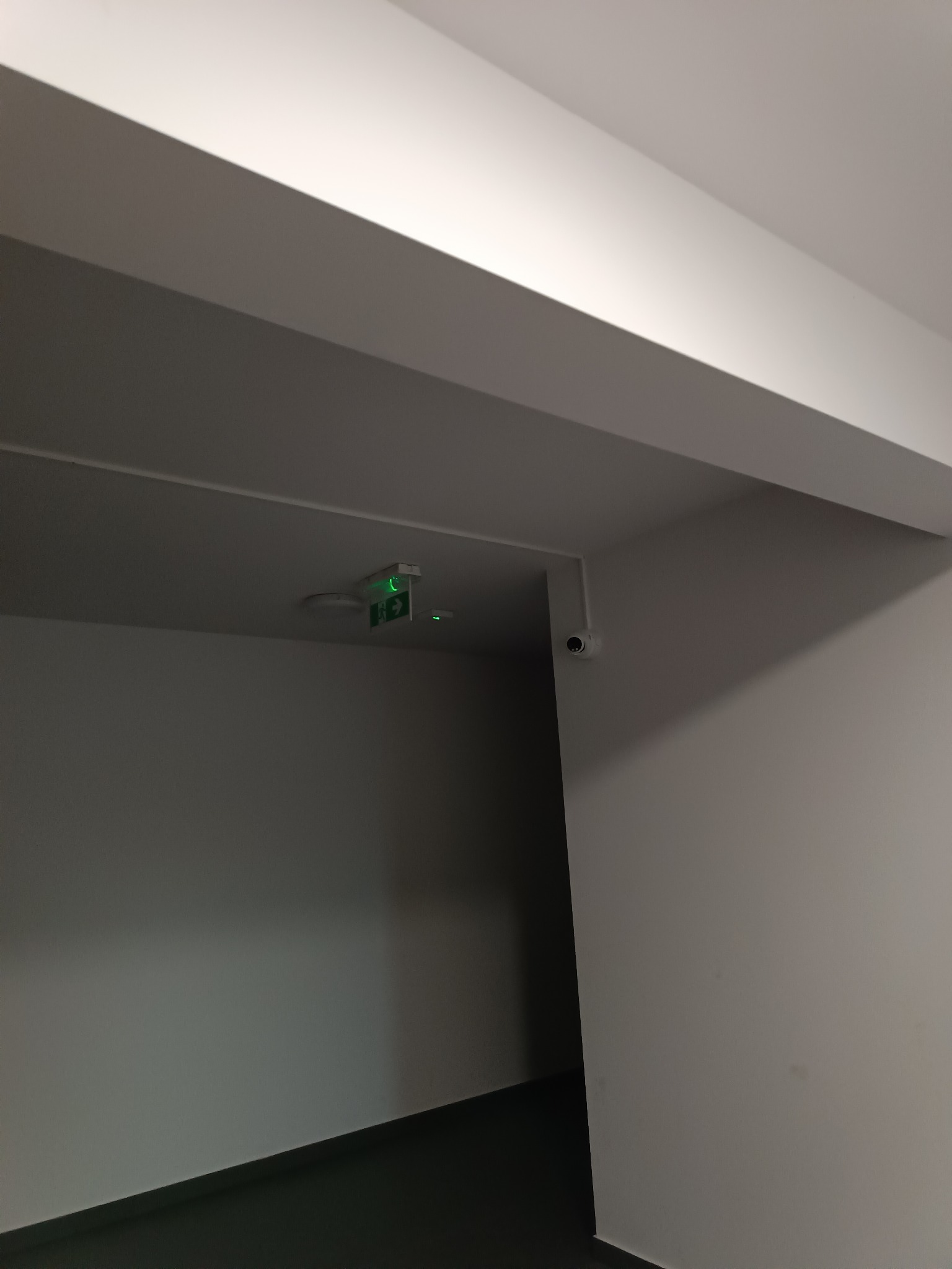 Kamera monitoringu kopułkowa zamontowana na ścianie w rogu korytarza, obok oświetlenia awaryjnego z zielonym piktogramem drogi ewakuacyjnej.