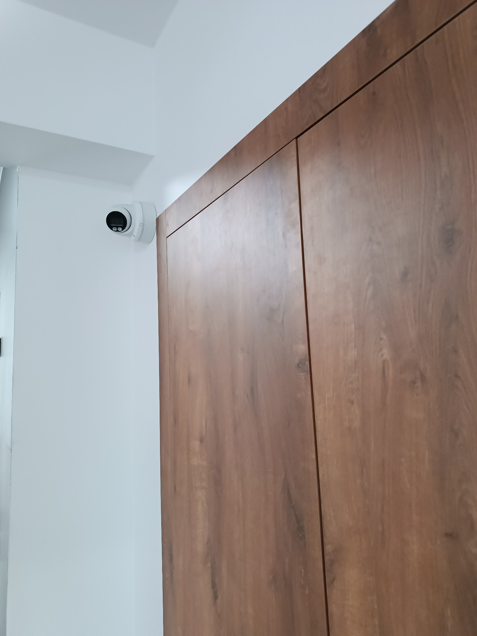 Kamera monitoringu kopułkowa, biała, zamontowana na białej ścianie obok drewnianej szafy wnękowej.
