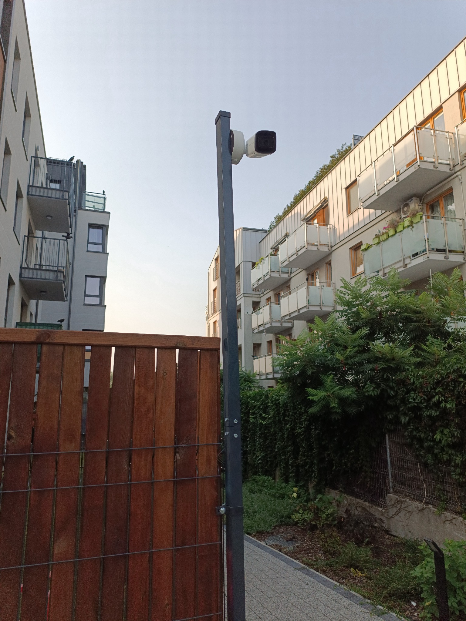 Zewnętrzna kamera monitoringu na słupie, widok na nowoczesne budynki mieszkalne z balkonami i drewniane ogrodzenie.