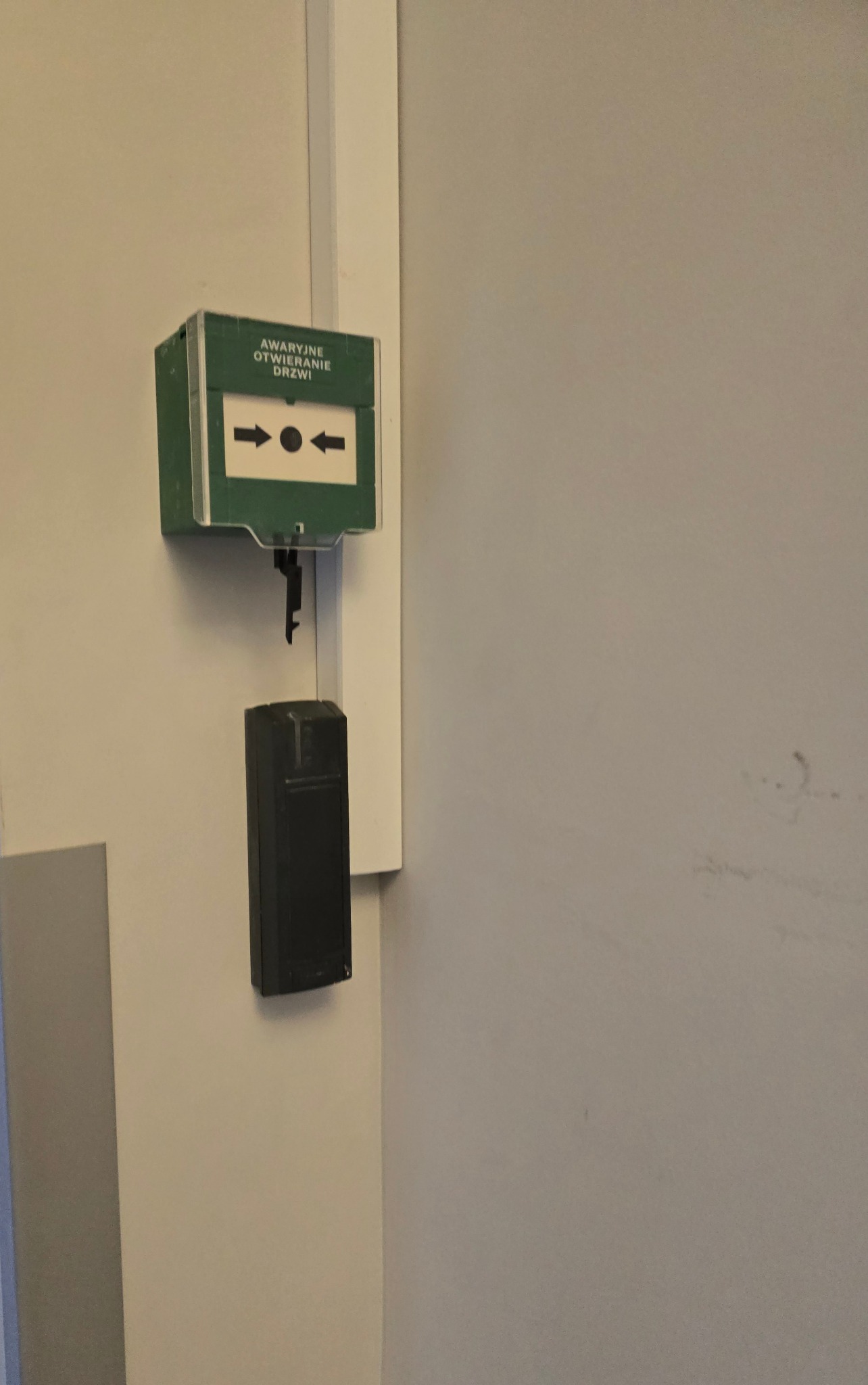 Zielona obudowa z napisem 'Awaryjne Otwieranie Drzwi' i strzałkami, pod nią czarny moduł z kluczem do awaryjnego otwierania drzwi, zamontowane na białej ścianie.