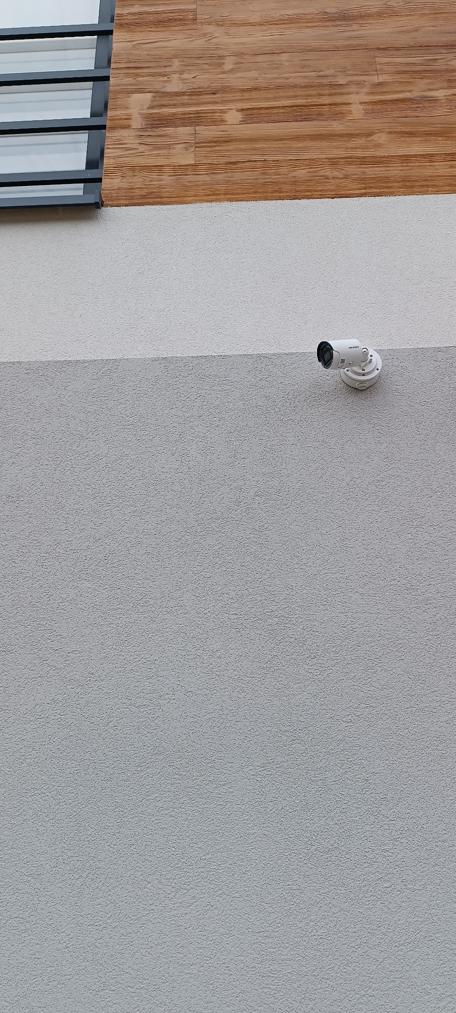 Biała kamera monitoringu zamontowana na szarej, teksturowanej ścianie budynku, z fragmentem drewnianej okładziny i okna w górnej części kadru.