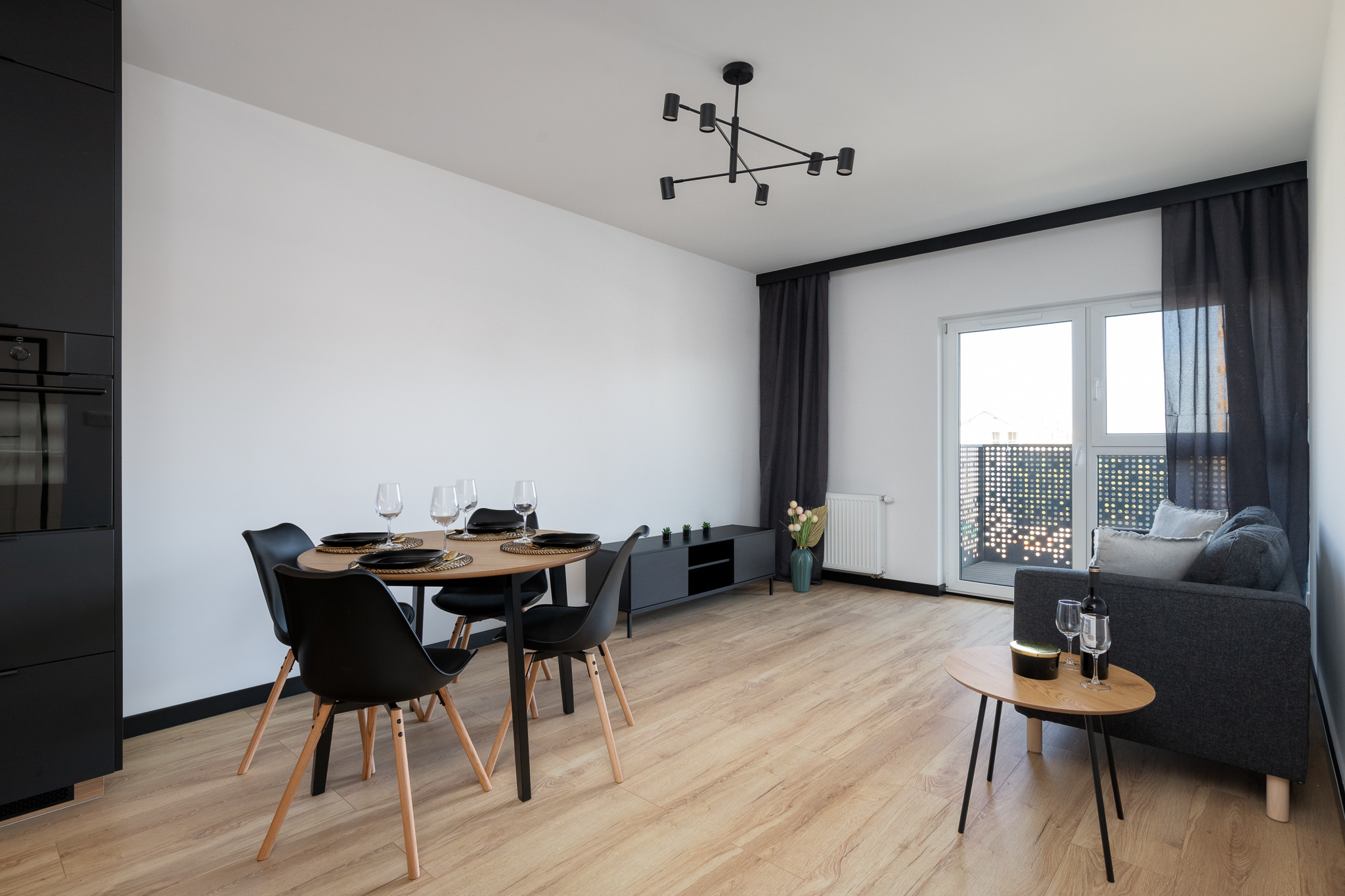 Jasne wnętrze mieszkania w minimalistycznym stylu: stół z czarnymi krzesłami i nakryciami, szklanki do wina, sofa z poduszkami, drewniana podłoga, czarne akcenty w postaci karniszy i szafek...