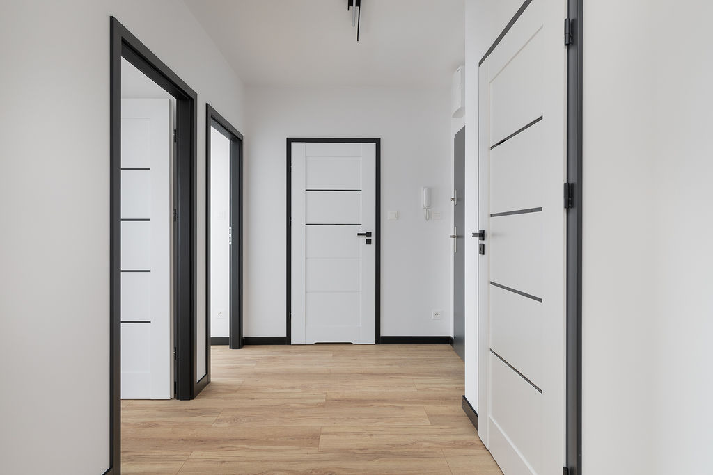 Białe drzwi z czarnymi akcentami w jasnym korytarzu z panelami podłogowymi imitującymi drewno.