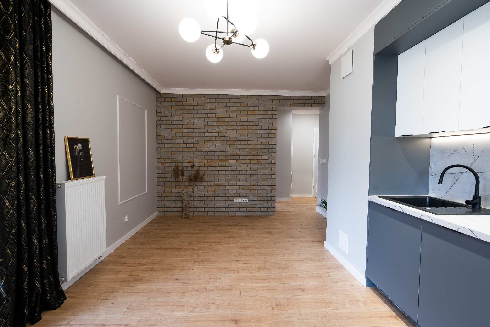 Jasne, przestronne wnętrze po remoncie z elementami dekoracyjnymi: ściana z cegły, nowoczesna lampa sufitowa, ciemne zasłony i szara zabudowa kuchenna z marmurkowym blatem.