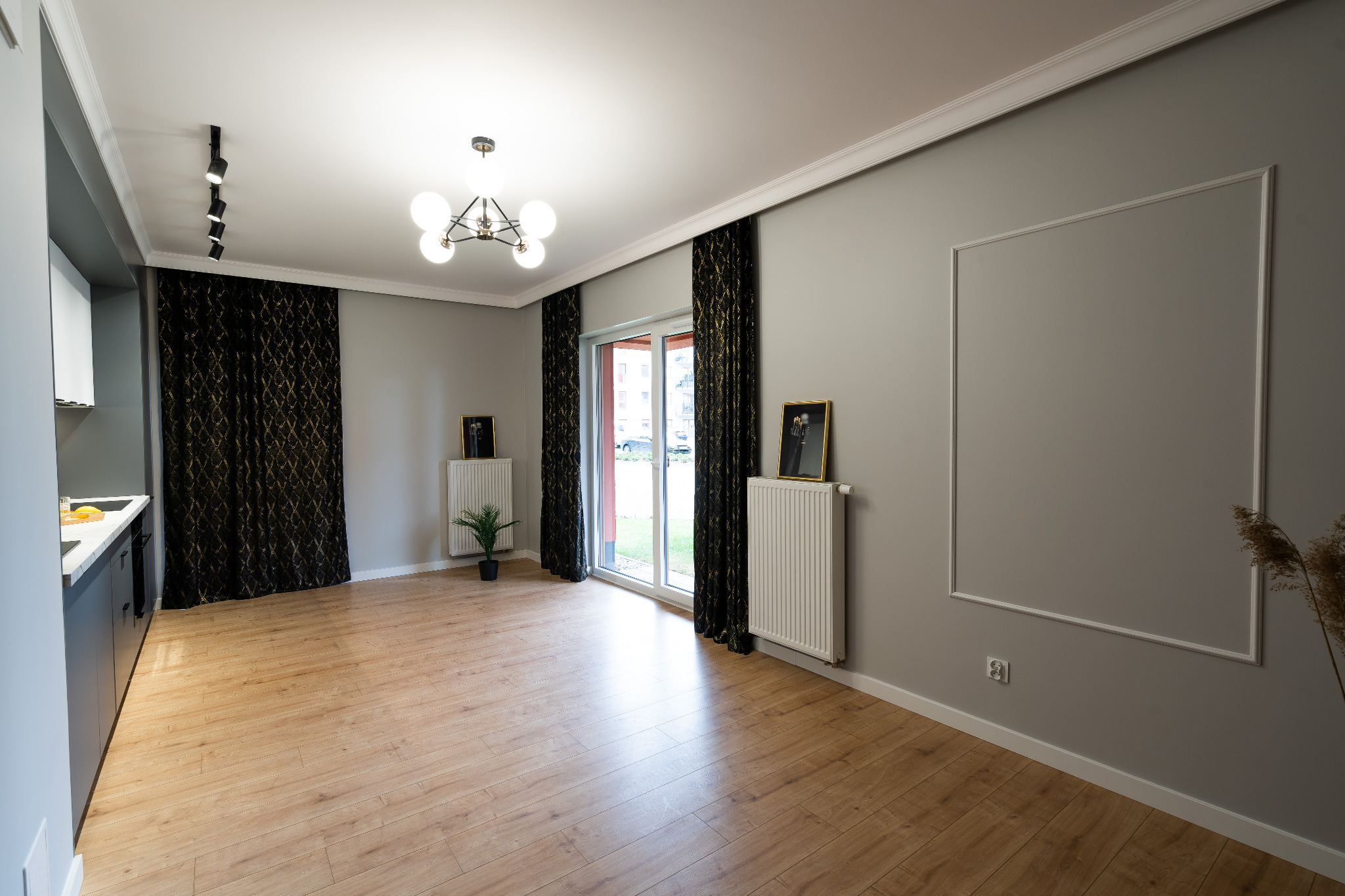 Jasne wnętrze pokoju z szarą ścianą, drewnianą podłogą, białymi grzejnikami i czarnymi zasłonami ze złotym wzorem, widok na okno balkonowe.