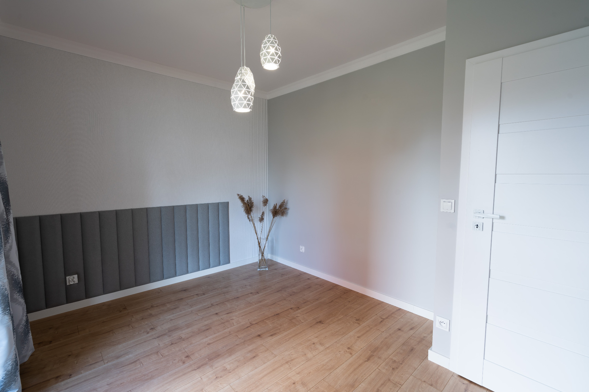 Jasne, puste pomieszczenie z drewnianą podłogą, szarą tapetą w paski, dekoracyjnym panelem ściennym z tkaniny, białymi listwami przypodłogowymi i białymi drzwiami, oświetlone przez dwie...
