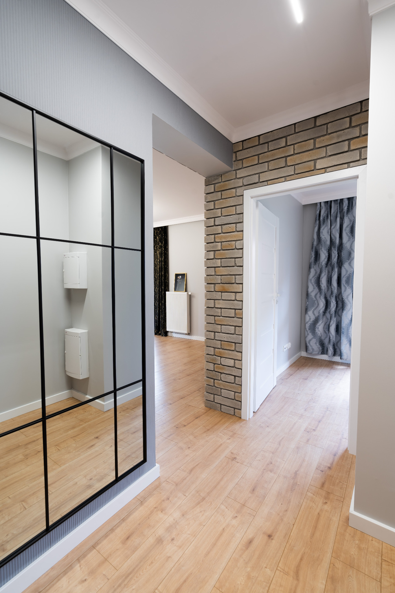 Hallway interior featuring a large, grid-patterned mirror reflecting white electrical boxes, contrasting with a brick-clad doorway and light wood flooring.