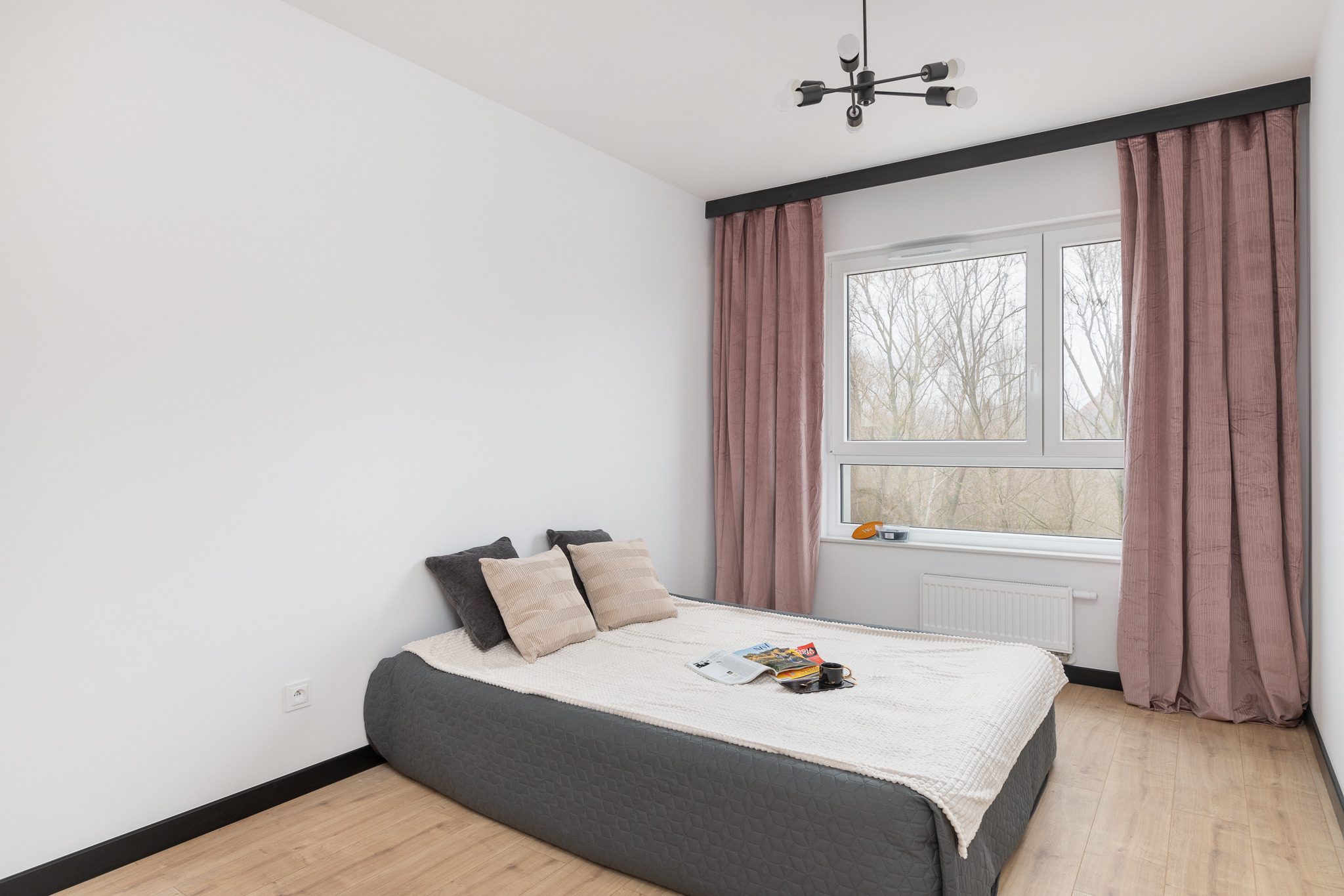 Minimalistyczna sypialnia z łóżkiem kontynentalnym w szarej tapicerce, jasną pościelą, poduszkami w odcieniach beżu i szarości, oraz czarnym żyrandolem pająkiem.