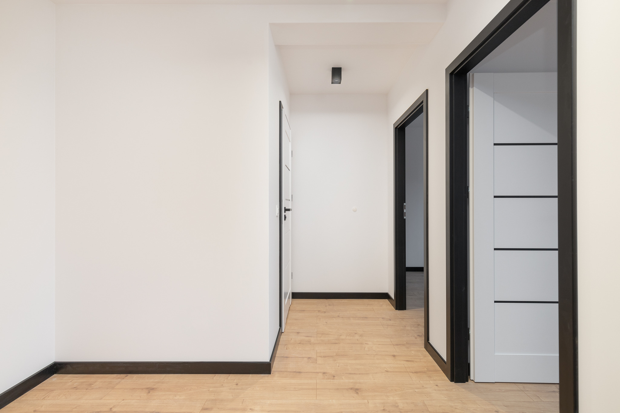 Jasne wnętrze mieszkania po remoncie: białe ściany, drewniana podłoga, czarne listwy przypodłogowe i ościeżnice drzwiowe.