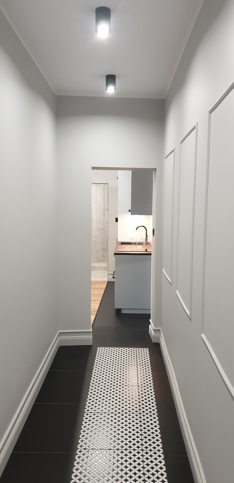 Wnętrze mieszkania: korytarz z czarną podłogą, białymi ścianami z dekoracyjnymi listwami i widokiem na kuchnię z czarną armaturą i drewnianym blatem.