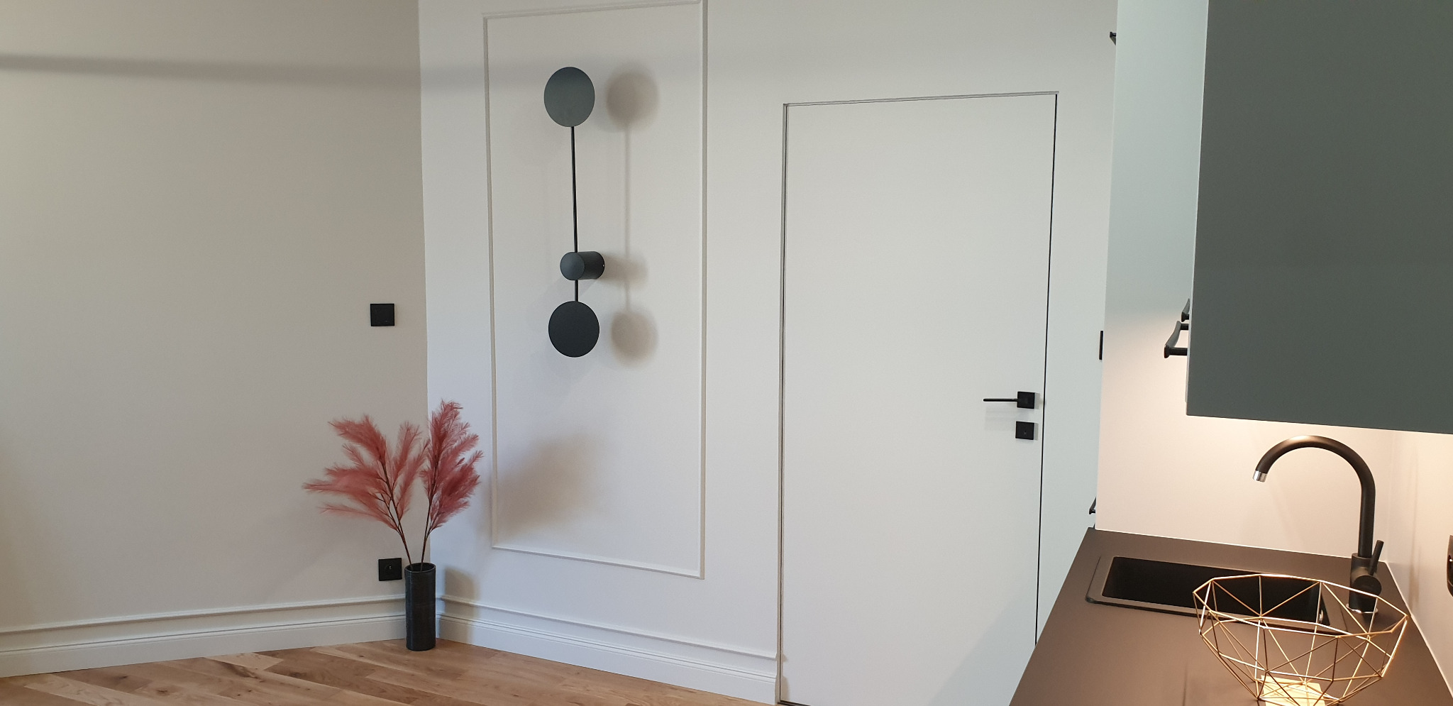 Minimalistyczne wnętrze: białe ściany z dekoracyjną sztukaterią i nowoczesną czarną ozdobą ścienną, różowe trawy pampasowe w ciemnym wazonie, białe drzwi z czarną klamką oraz fragment szarej szafki...