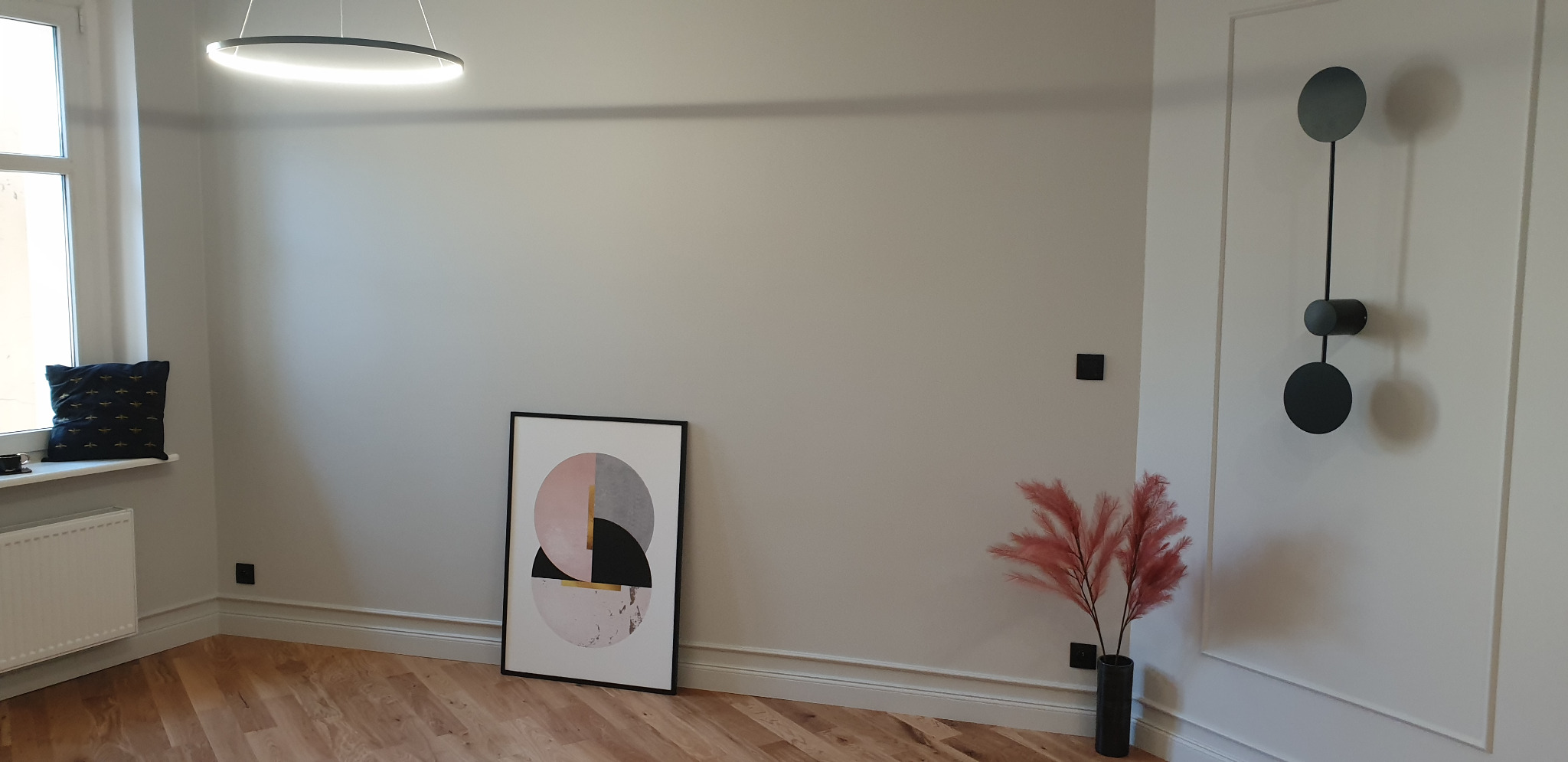 Jasne wnętrze po odświeżeniu, z drewnianą podłogą, minimalistycznym oświetleniem i dekoracjami ściennymi: abstrakcyjny obraz w czarnej ramie i metalowa ozdoba z okręgów.