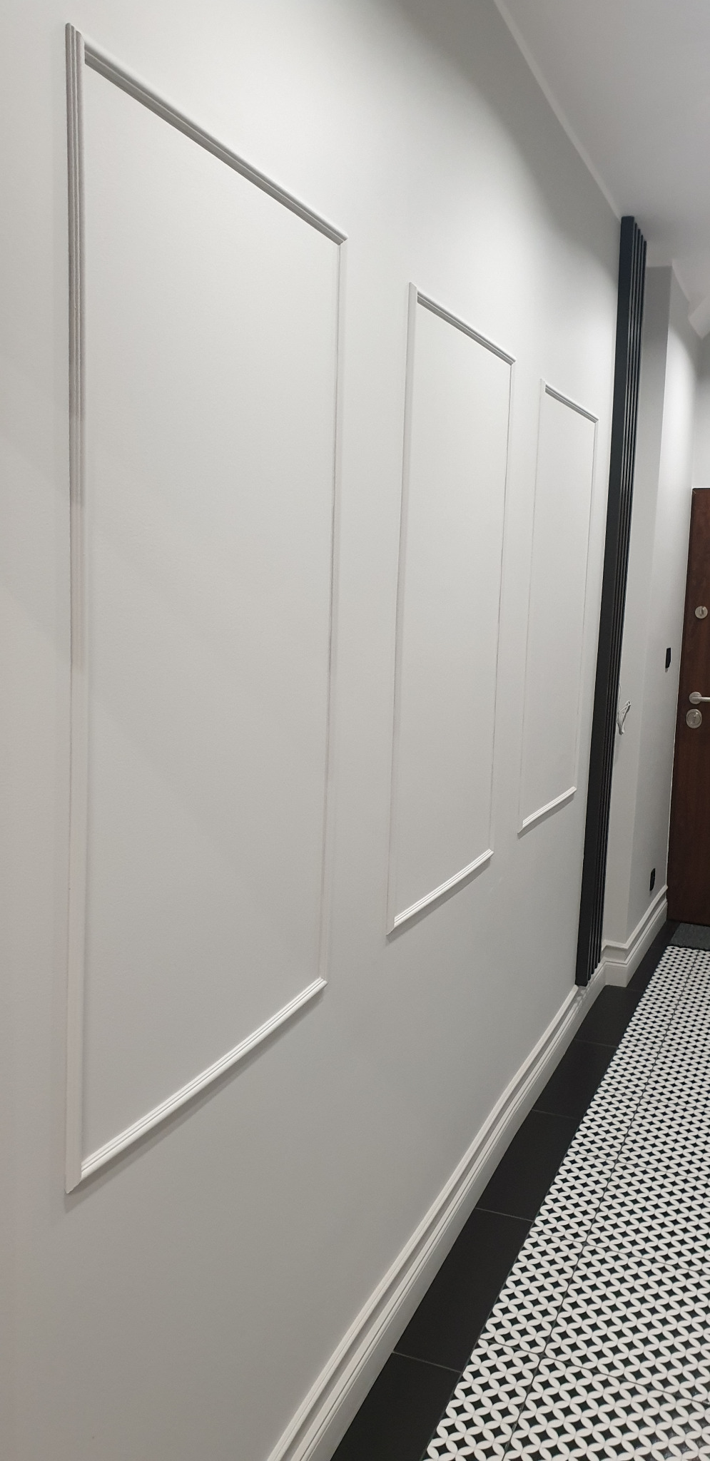 Ściana w jasnym odcieniu szarości z trzema prostokątnymi panelami dekoracyjnymi z białymi obramowaniami, czarna listwa grzejnikowa i podłoga wyłożona czarno-białą mozaiką.