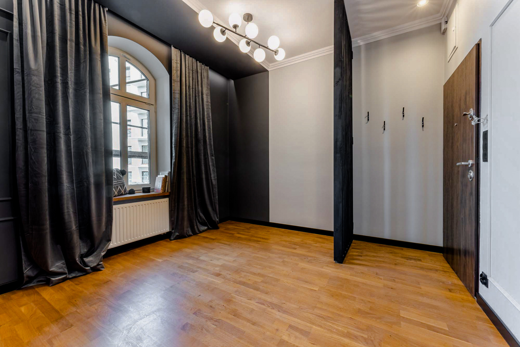 Jasny, przestronny pokój po remoncie z drewnianą podłogą, dużym oknem z ciemnymi zasłonami i nowoczesnym oświetleniem sufitowym.