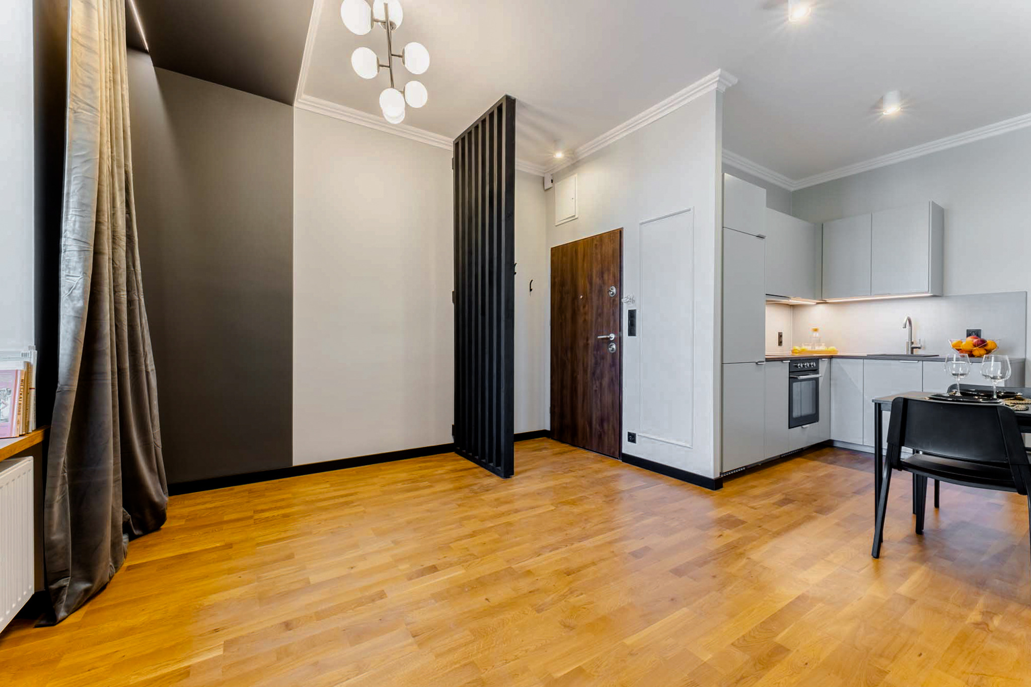 Jasny parkiet w nowoczesnym mieszkaniu z szarą ścianą, drewnianymi drzwiami wejściowymi i minimalistyczną kuchnią w tle.