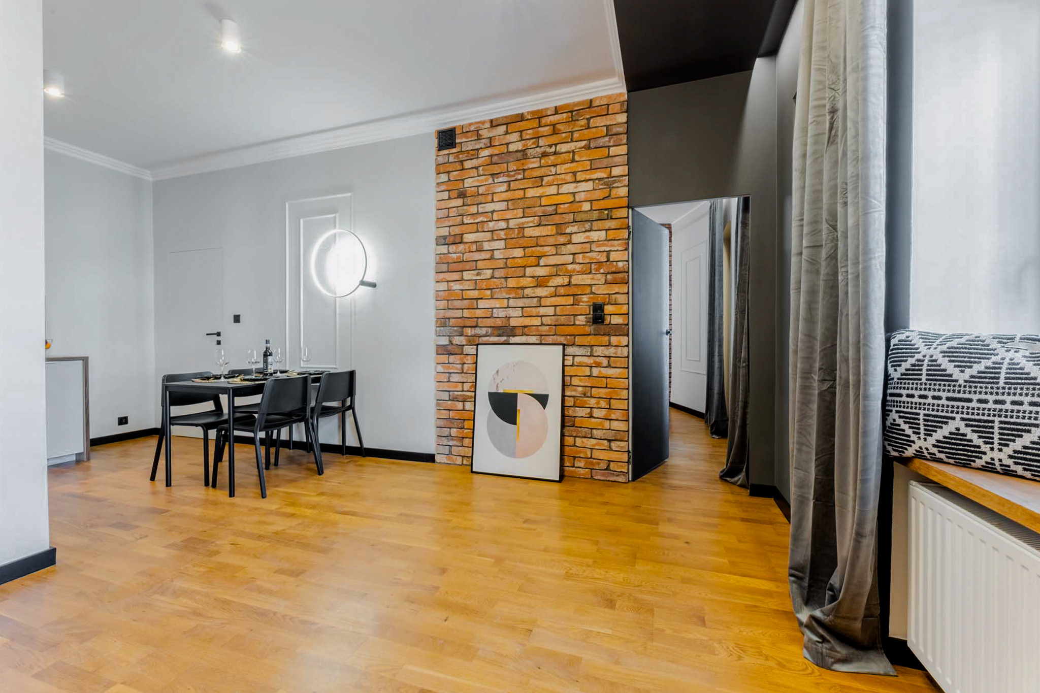 Jasne wnętrze mieszkania z parkietem, ceglaną ścianą, czarnym stołem z zastawą i minimalistycznym oświetleniem.