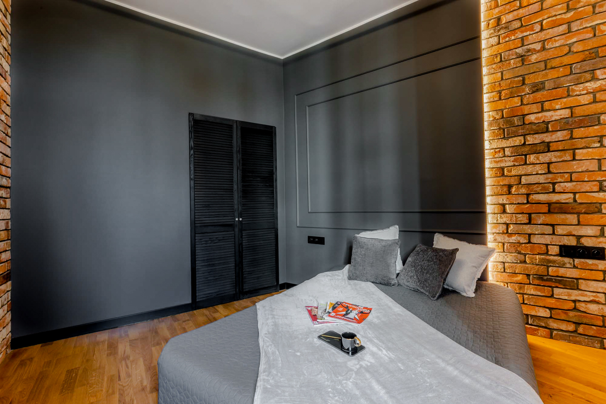 Sypialnia z szarą ścianą, ceglaną wstawką z podświetleniem, czarnym, żaluzjowym dwuskrzydłowym szafą i drewnianą podłogą; na łóżku leżą poduszki, gazety i czarna filiżanka na tacy.