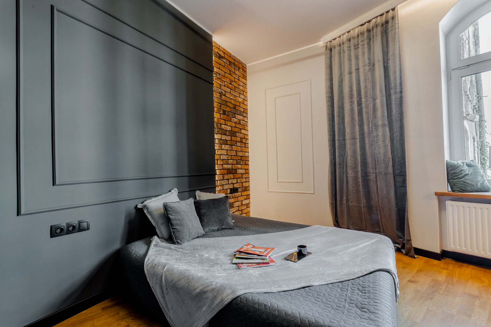 Pokój z pomalowaną na szaro ścianą z dekoracyjnymi listwami, fragmentem ściany z cegły, zasłoną i drewnianą podłogą.