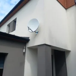 Biała antena satelitarna zamontowana na elewacji nowoczesnego domu z teksturą, widok z dołu, błękitne niebo w tle.