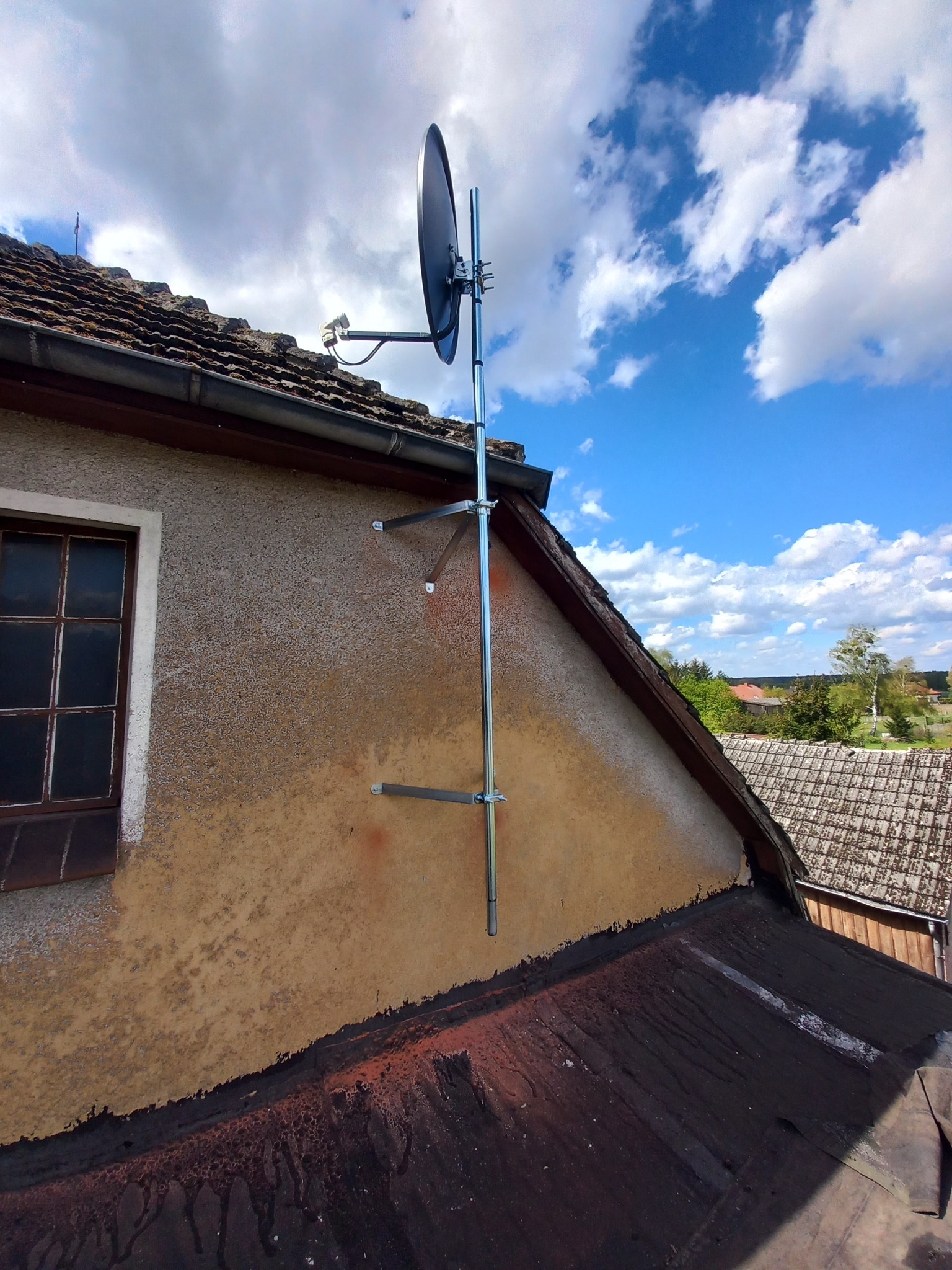Antena satelitarna zamontowana na metalowym maszcie przymocowanym do ściany budynku z oknem, na tle błękitnego nieba z chmurami.