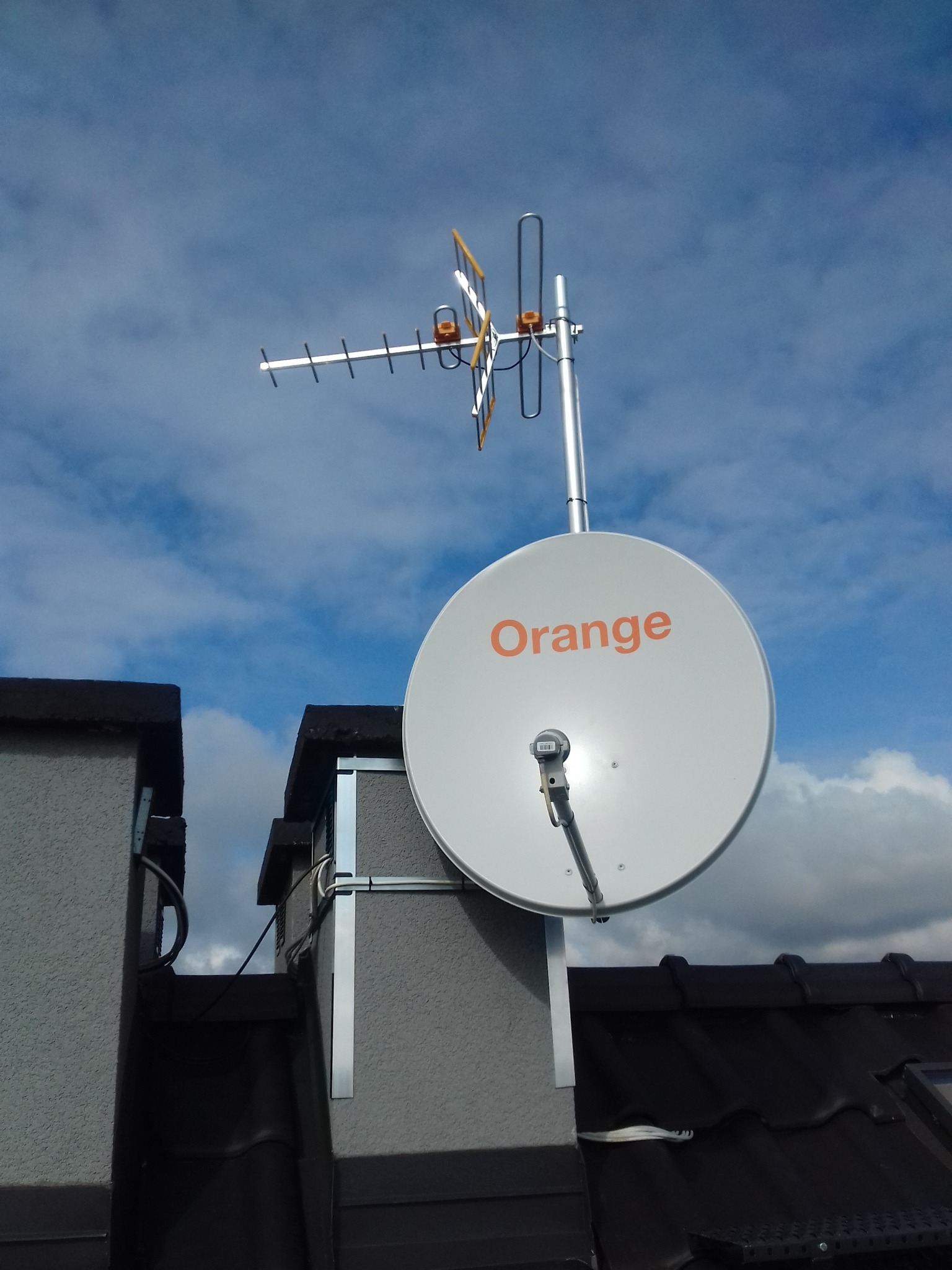 Antena satelitarna z logo Orange oraz antena naziemna zamontowane na dachu budynku, widoczne przewody i elementy mocujące, tło stanowi błękitne niebo z chmurami.
