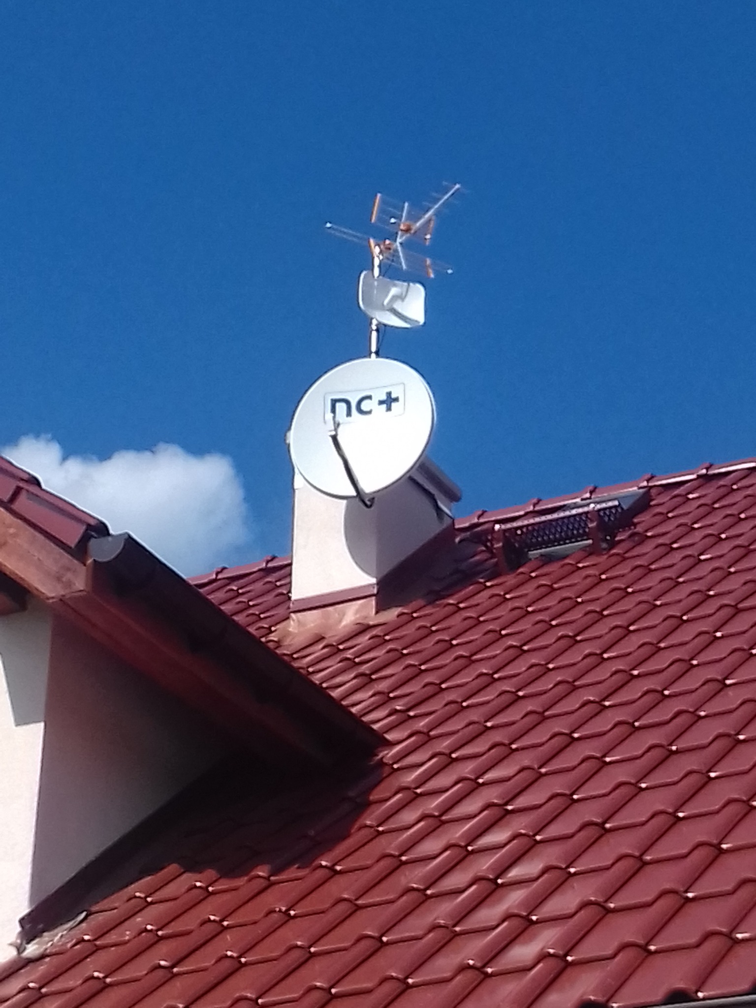 Antena satelitarna z logiem nc+ i antena naziemna zamontowane na kominie na dachu z czerwonej dachówki w słoneczny dzień.