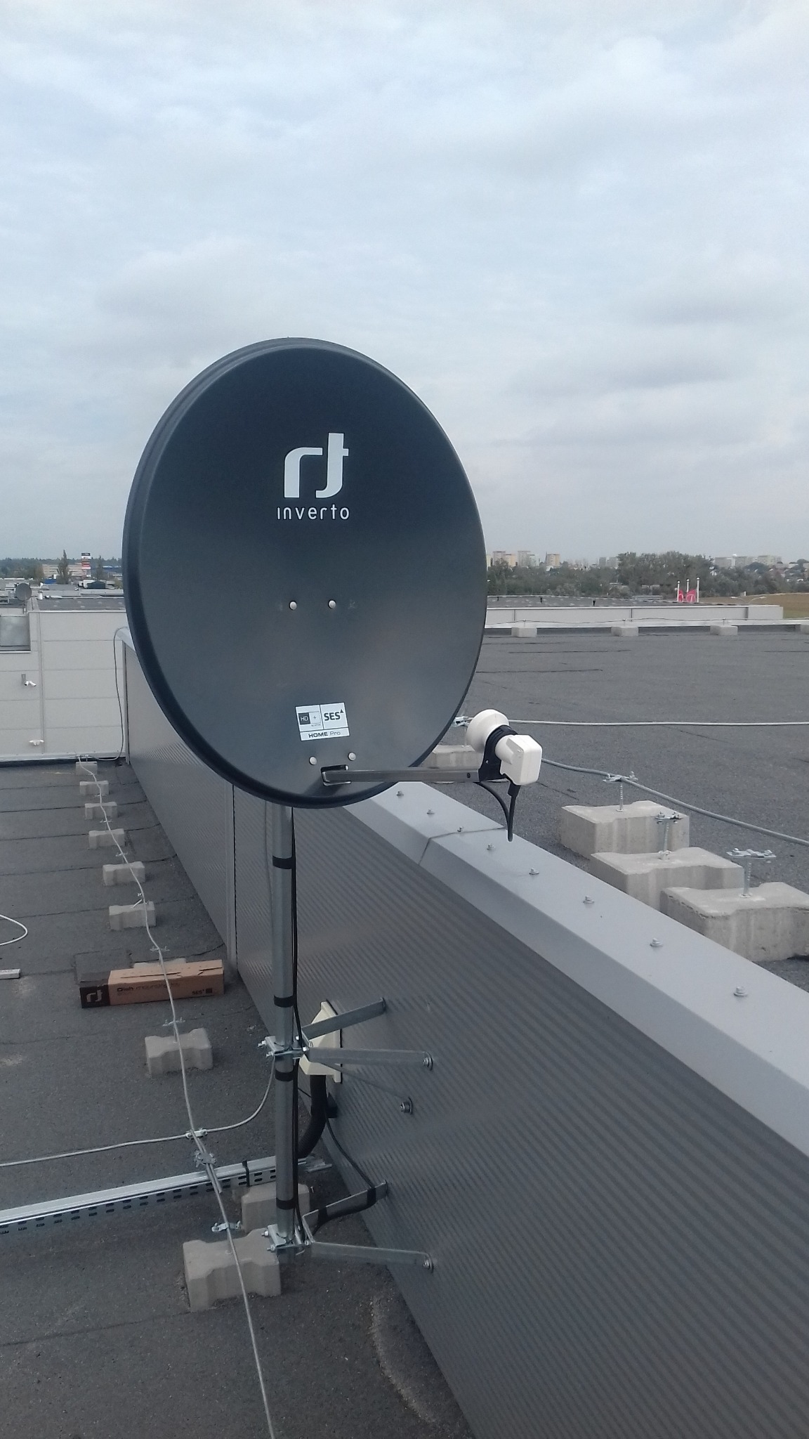 Czarna antena satelitarna marki Inverto zamontowana na płaskim dachu budynku, widoczne mocowania do ściany i przewody.
