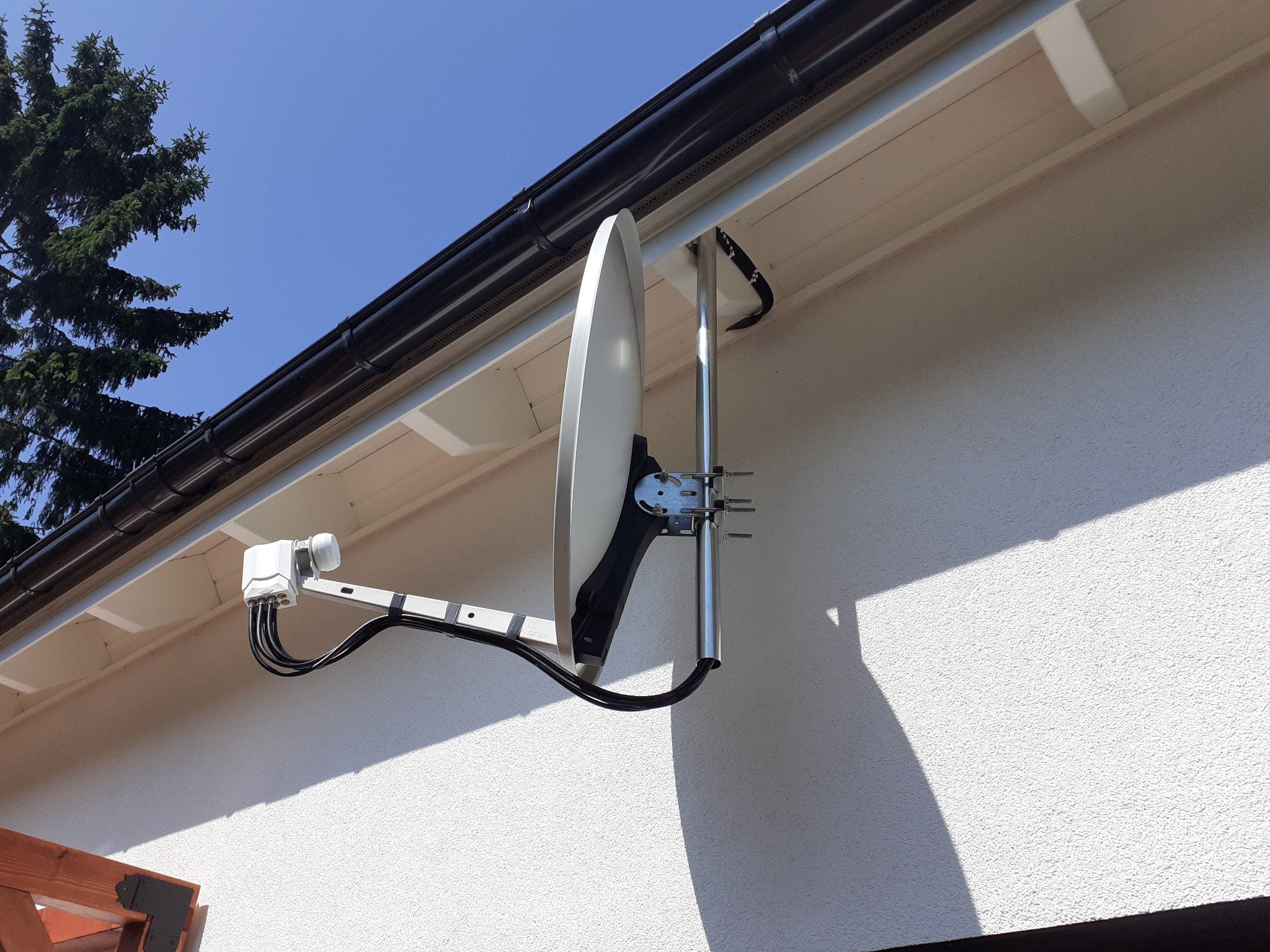 Biała antena satelitarna zamontowana na zewnętrznej ścianie budynku, widoczne kable i uchwyt montażowy, na tle błękitnego nieba i fragmentu dachu.