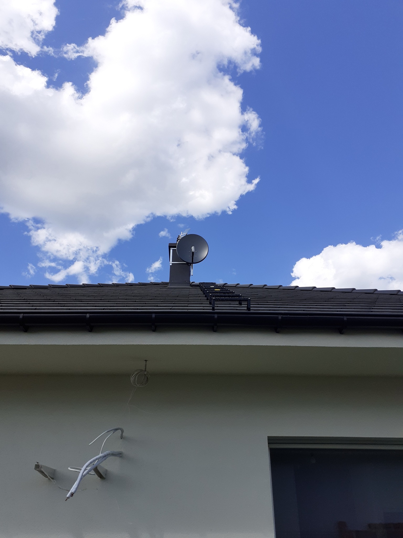 Antena satelitarna zamontowana na kominie na tle błękitnego nieba z obłokami, widoczne przewody na elewacji budynku.
